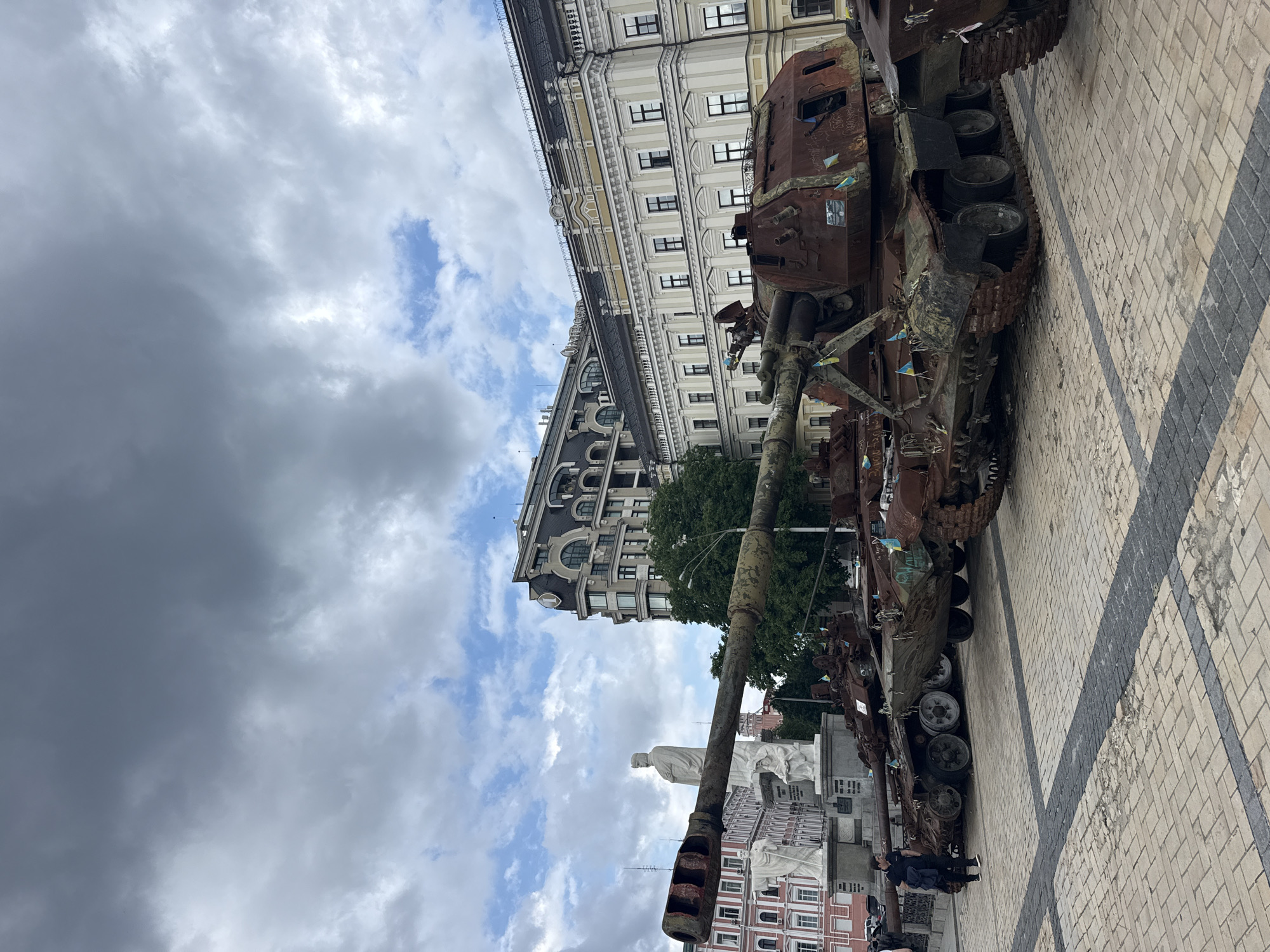 In Kyiv, Ukraine, a row of burnt-out tanks, some adorned with small Ukrainian flags, are displayed on a cobblestone plaza. The tanks, which appear to be military vehicles damaged in conflict, are positioned in front of historic buildings with ornate architecture and a statue of a figure in classical attire. The sky above is overcast with dramatic clouds. A lone individual in dark clothing is walking across the plaza, with their back to the camera. The scene suggests a public display of war remnants.