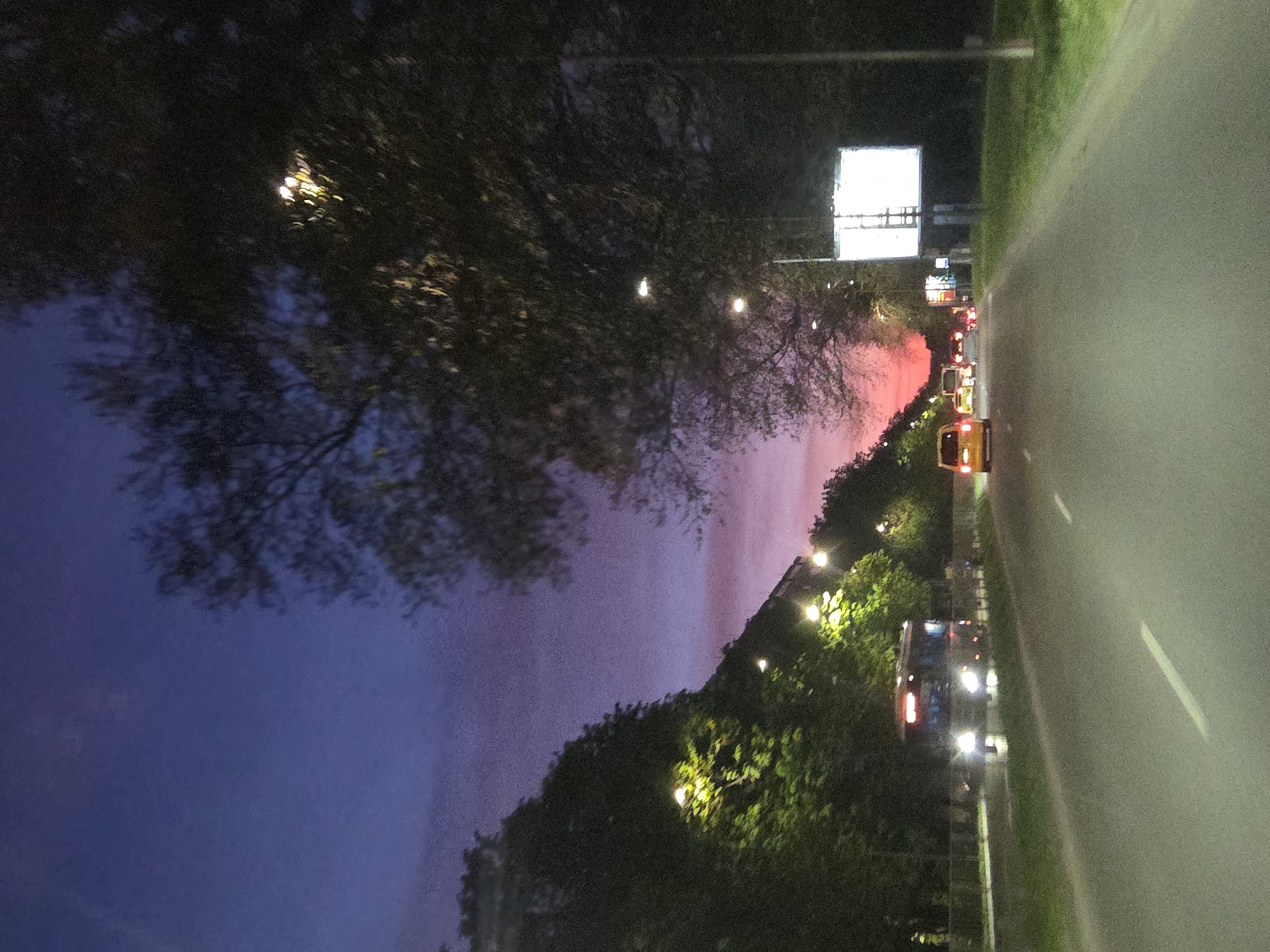 This image captures an evening or night scene of a multi-lane road in Varna, Bulgaria, lined with dense trees and illuminated by streetlights. The sky displays a striking gradient, transitioning from a deep blue-purple on the left to a vibrant pinkish-red on the right, possibly indicative of a late sunset or significant urban light pollution.
On the right side of the frame, the asphalt road stretches into the distance, marked by a clear white dashed line separating the lanes. Several vehicles are visible on the road, their headlights and taillights brightly lit, suggesting ongoing traffic. A distinct yellow vehicle, possibly a taxi or a small van, stands out in the middle distance, while a larger vehicle, perhaps a bus or truck, is visible closer to the foreground.
Both sides of the road are densely covered with trees and foliage, some branches appearing bare against the colorful sky. Streetlights cast bright glows on the leaves and illuminate portions of the road. In the distance, on the right side of the road, a large, bright white billboard or sign structure stands out, though no specific text or imagery is legible on it. Smaller, indistinct signs are also present further down the road. The scene suggests a clear evening with active road traffic. No individuals are visibly present.