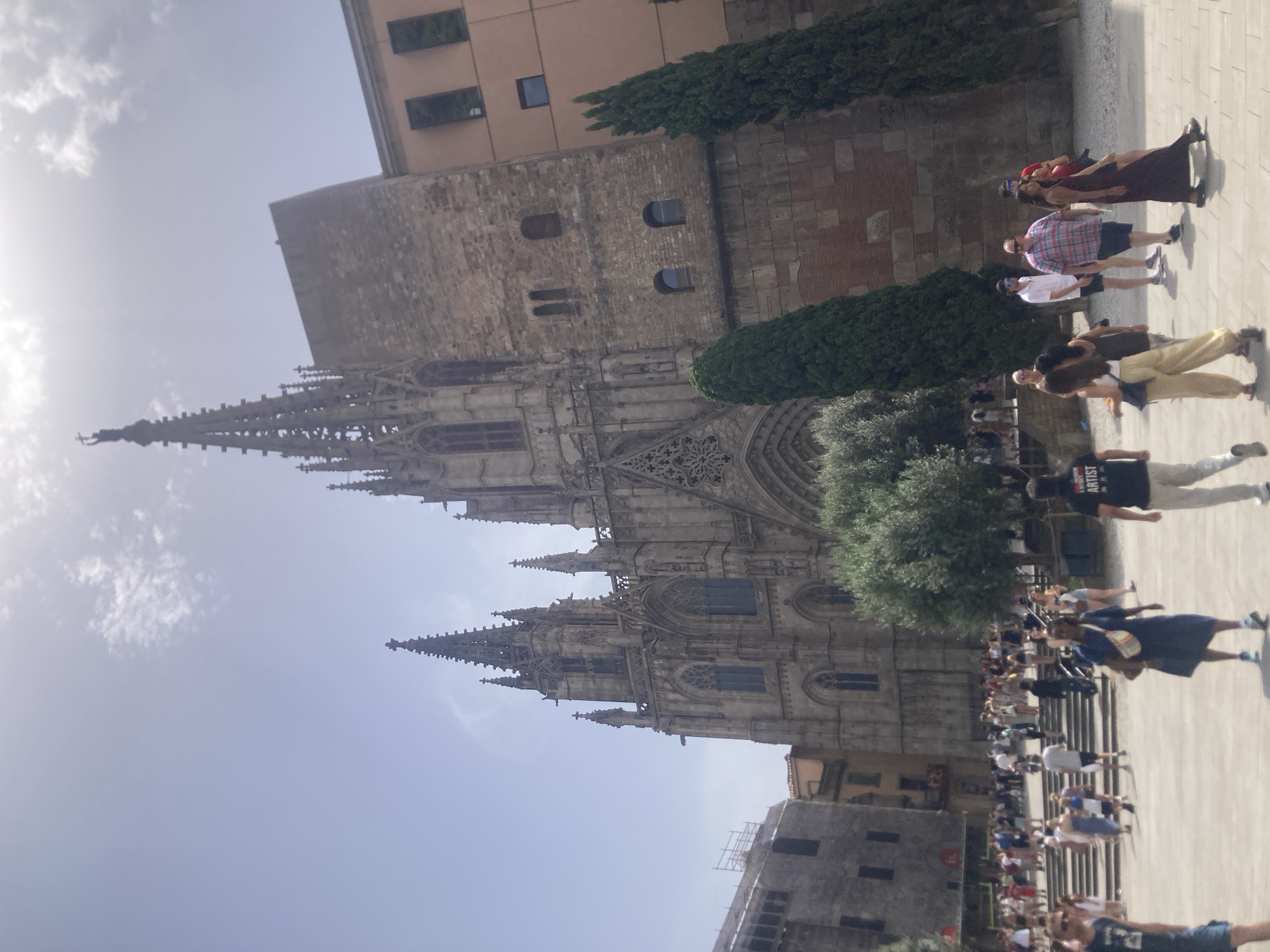 The image depicts a sunny day in Barcelona, Spain, showcasing the Barcelona Cathedral. In the foreground, a crowd of tourists is visible, dressed in casual summer attire. People are walking in various directions, some standing and observing the architecture. To the left, steps lead up to the cathedral, with more people ascending. The Barcelona Cathedral, a Gothic structure, dominates the background with its intricate spires and stone facade. Adjacent to the cathedral, a modern building with a pink facade and dark windows is situated. Tall, dark green cypress trees stand between the buildings and in front of the cathedral. The sky is bright blue with scattered white clouds.