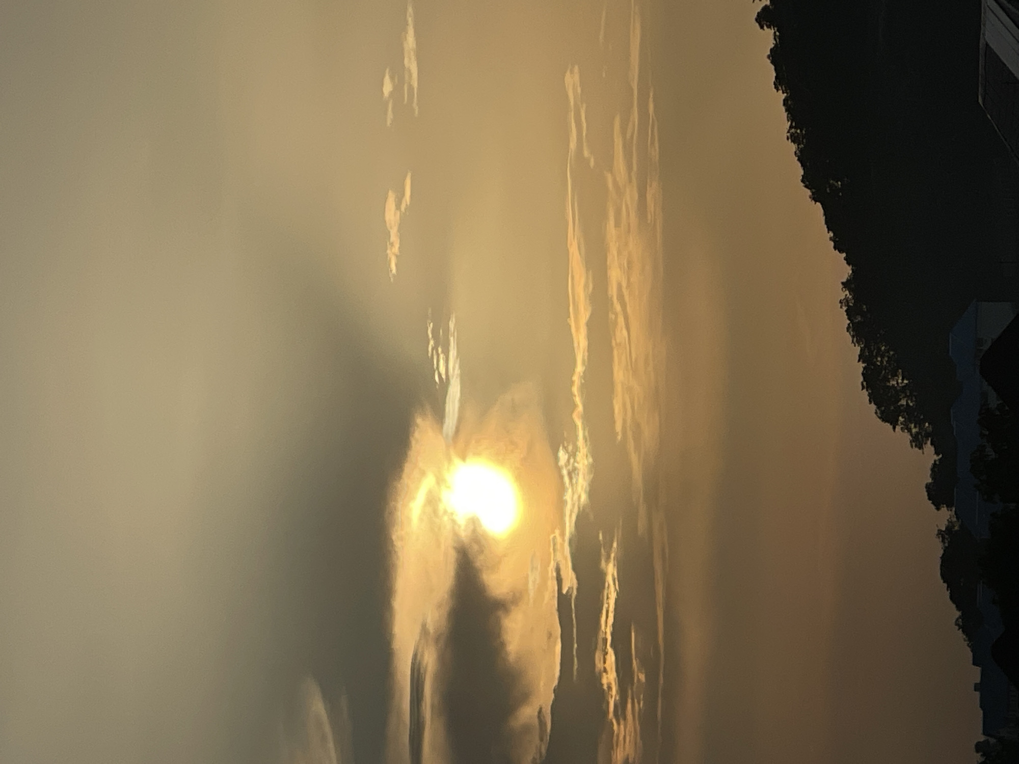 This is a photograph taken at sunset in Abuja, Nigeria. The sun, a bright orb of light, is partially obscured by clouds, casting a warm, golden glow across the sky. The clouds themselves vary in texture and density, with some appearing wispy and translucent, while others are more opaque and shadowed. Below the sky, a silhouetted hillside is visible, covered in a dense growth of trees. In the foreground, the tops of buildings and rooftops can be seen, also in silhouette against the fading light. The overall impression is one of natural beauty and the tranquil end of the day in a Nigerian city.