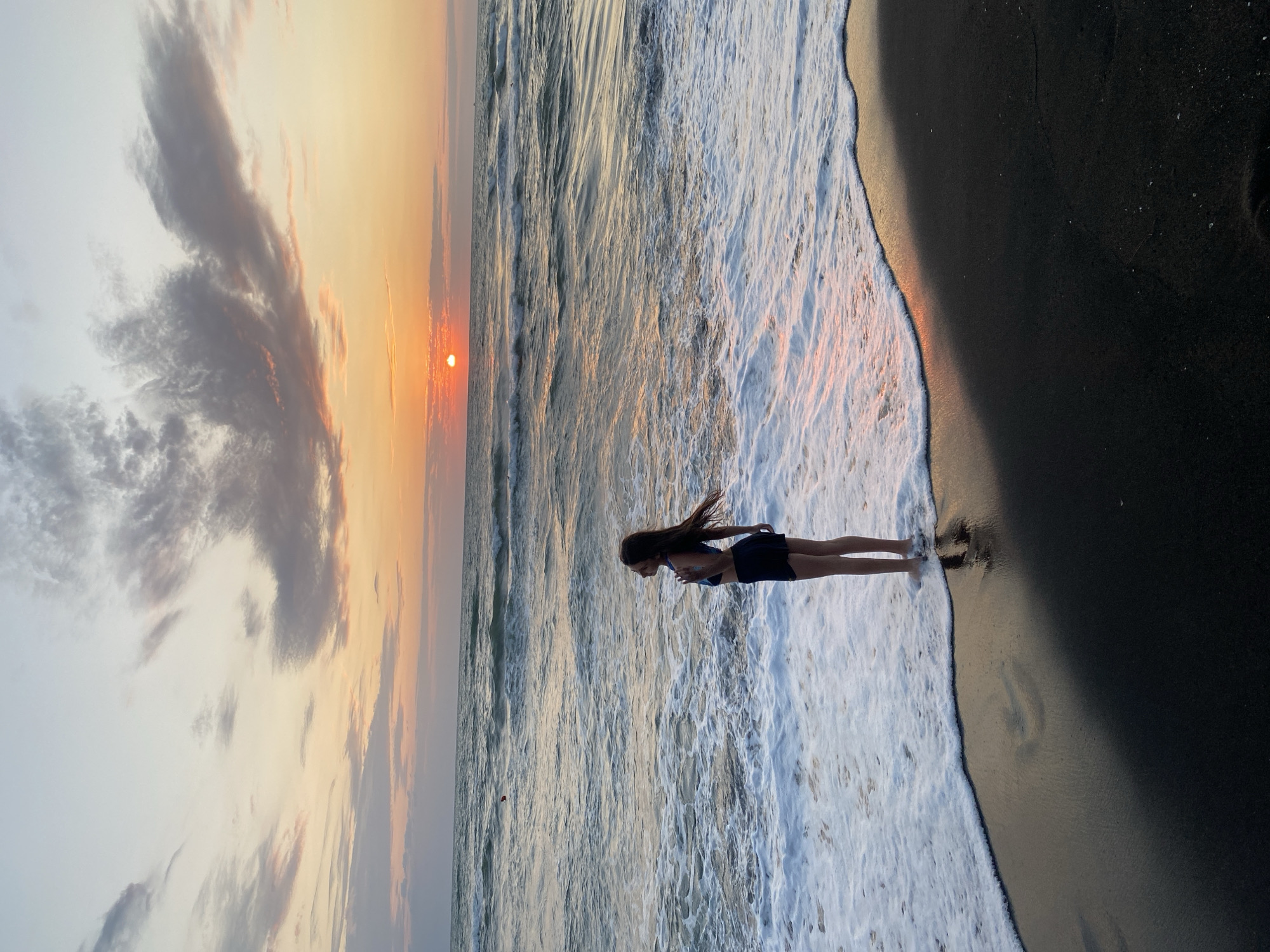 A young girl stands in the shallow water on a beach at sunset. The sun is low on the horizon, casting a warm glow on the clouds and water. The girl is facing away from the camera, looking out at the ocean. Her hair is blowing in the wind. The beach is dark, possibly black sand, and the waves are crashing around her feet. The sky is filled with soft, pastel-colored clouds. The overall scene is peaceful and serene.