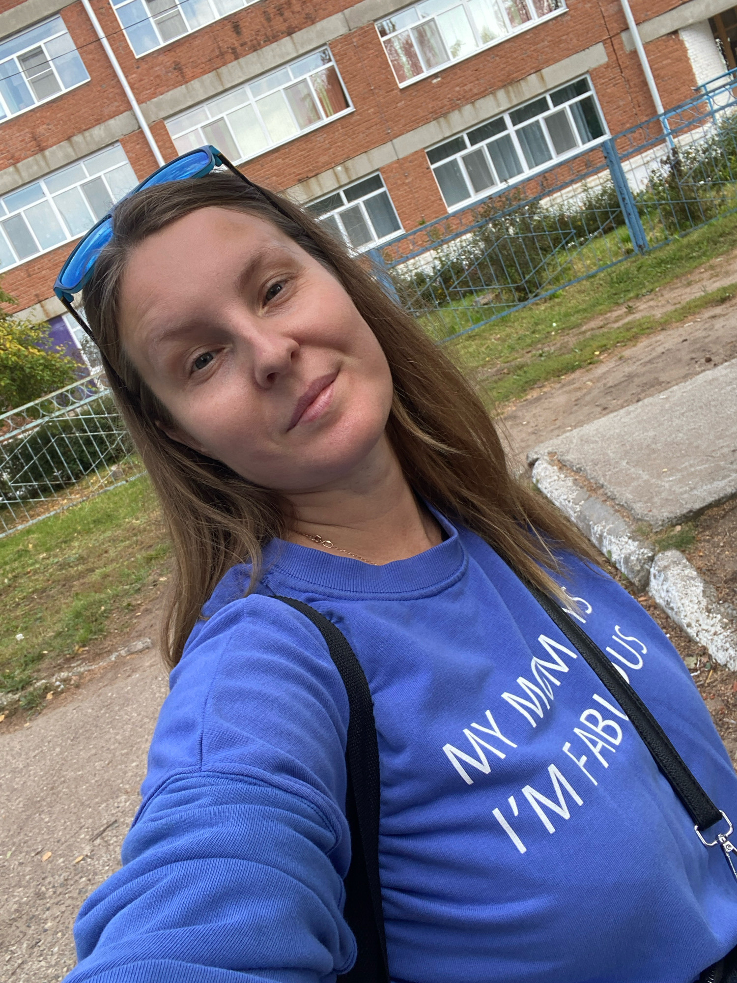 A woman with long brown hair and blue sunglasses perched on her head takes a selfie outdoors. She is wearing a bright blue sweatshirt with white text that reads, "MY MOM IS FABULOUS." A black strap from a bag is visible across her shoulder. The background features a red brick building with multiple windows and a blue metal fence. Green grass and dirt are visible in the foreground, suggesting an outdoor, possibly urban or suburban, setting. The lighting indicates it is daytime, and the weather appears to be overcast. The woman's expression is relaxed and she is smiling slightly, suggesting a casual and pleasant moment. The scene is likely in Noviy, Russia, as per the location context.