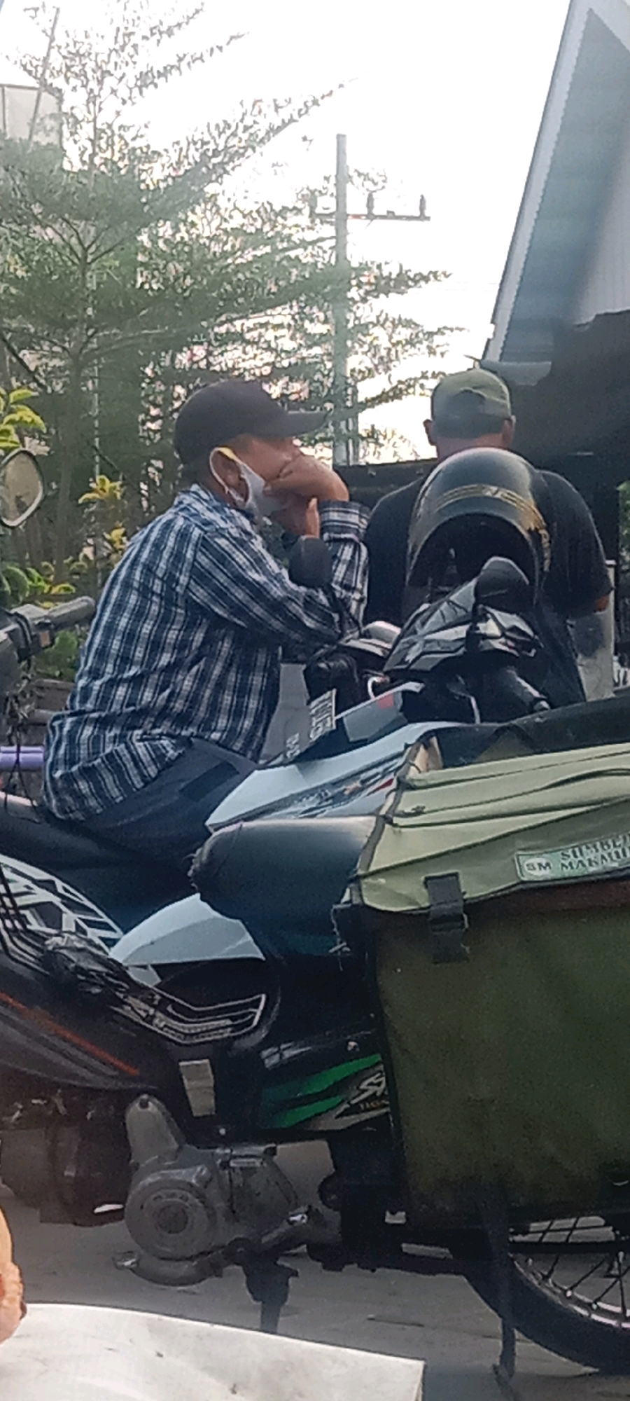 In Kras, Indonesia, two individuals are positioned on a motorcycle. The person in the foreground, wearing a checkered shirt and a black baseball cap, has a white face mask partially covering their face and is resting their chin on their hands. Their right hand is on their chin, and their left arm is bent, resting on their chest. The motorcycle itself is white and blue, with a black engine and chrome accents. A dark green bag is attached to the rear of the motorcycle, with a label reading "SM MAKAWU". Behind the first individual, another person is visible, wearing a green cap and a dark t-shirt. They are looking away from the camera. The scene appears to be outdoors, with trees and a utility pole visible in the background under a bright sky, suggesting daylight. The overall setting is urban or suburban.