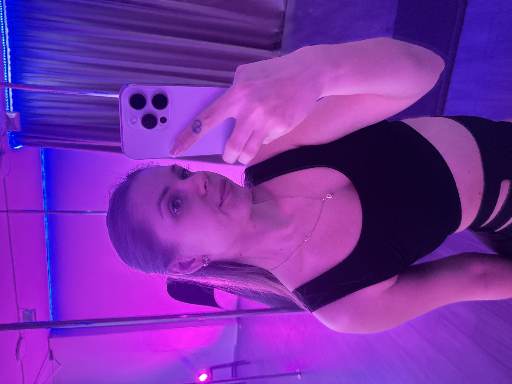 A woman is taking a selfie with her iPhone in a room with a blue and purple neon light strip. She is wearing a black top and has a tattoo of a clover on her index finger. Her blond hair is pulled back. The location is Batumi, Georgia.