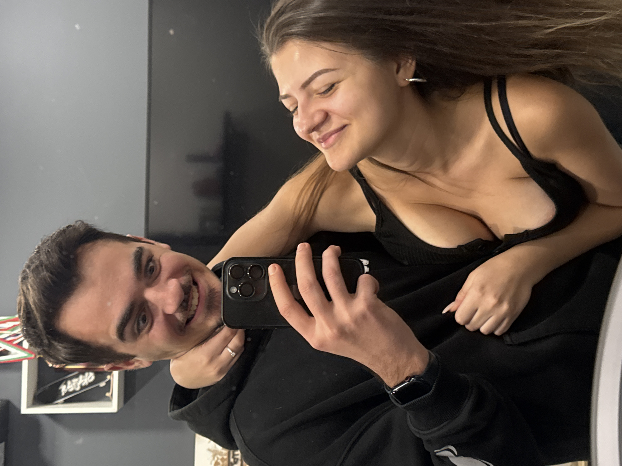 A man and a woman are posing for a selfie indoors. The man is holding a black smartphone and smiling, while the woman is also smiling and looking at him. She is wearing a black tank top. The man is wearing a black hooded sweatshirt and a smartwatch. In the background, there is a gray wall and a shelf with objects on it. The location is Sofia, Bulgaria.