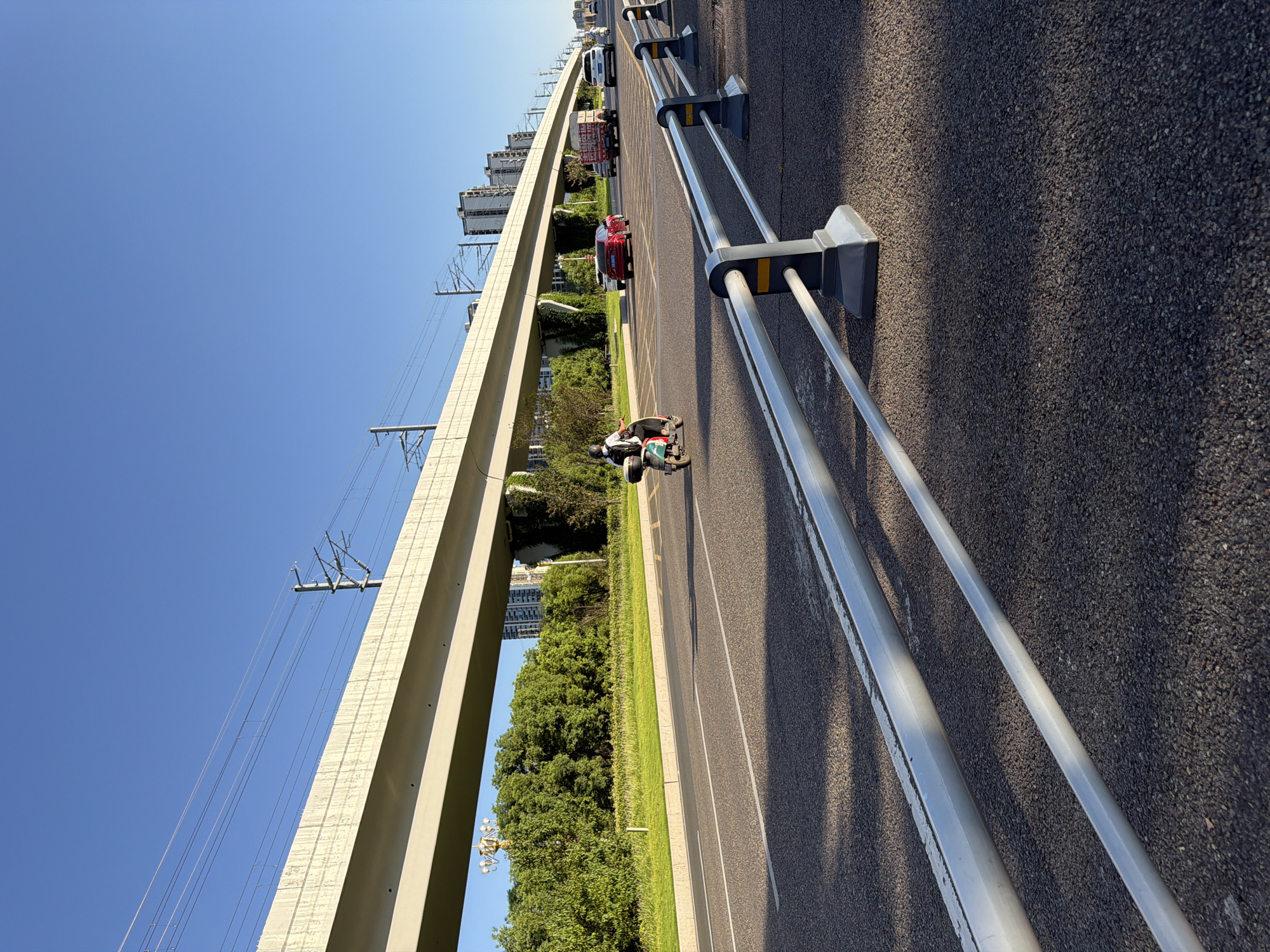 A scooter rider wearing a helmet and a backpack is traveling on a road in the city of Jiangjin, China, under a clear blue sky. The road has lane markings and is bordered by a metal guardrail. In the background, a large elevated structure, likely an overpass, runs parallel to the road, with power lines and utility poles visible. Beyond the overpass, there are rows of trees and modern buildings. Several cars, including a red sedan and a white SUV, are also on the road, moving in the same direction as the scooter. A truck with a red cargo box is also present. The sun is casting shadows on the road, indicating it is likely late afternoon or early morning.