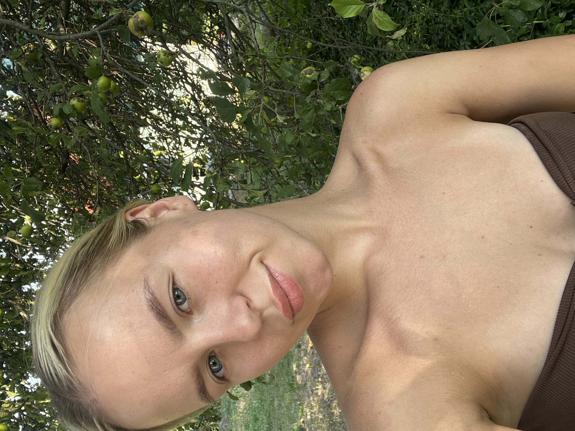 A medium-shot, outdoor selfie captures a young, fair-skinned woman from the chest up, her blond hair pulled back. She is positioned in front of a backdrop of lush green foliage, including a tree with small green apples. The woman is looking directly at the camera with a slight smile. Her shoulders and décolletage are exposed, and she appears to be wearing a brown strapless top. The lighting suggests it is daytime, with sunlight casting shadows and highlighting her features. The background is softly focused, emphasizing the woman as the primary subject.