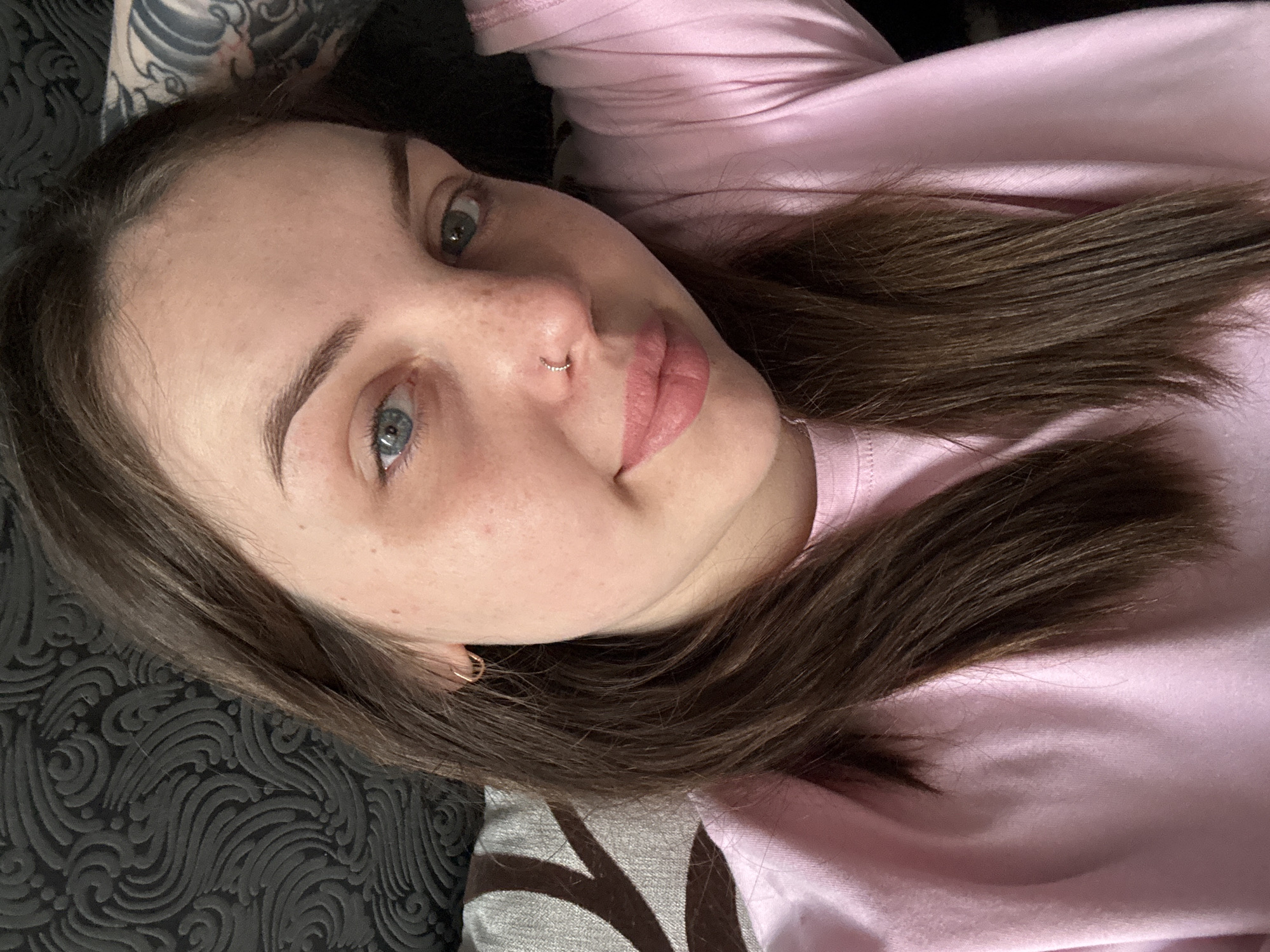 A young woman with light skin, blue eyes, and brown hair is pictured. She is wearing a pink shirt and has a nose ring in her left nostril and a small hoop earring in her left ear. She appears to be lying on a patterned gray and black surface, possibly a couch or bed. There is a tattoo visible on her left arm. The image does not show anything specific to locate this image in the city of Chornomorskoe, Ukraine.