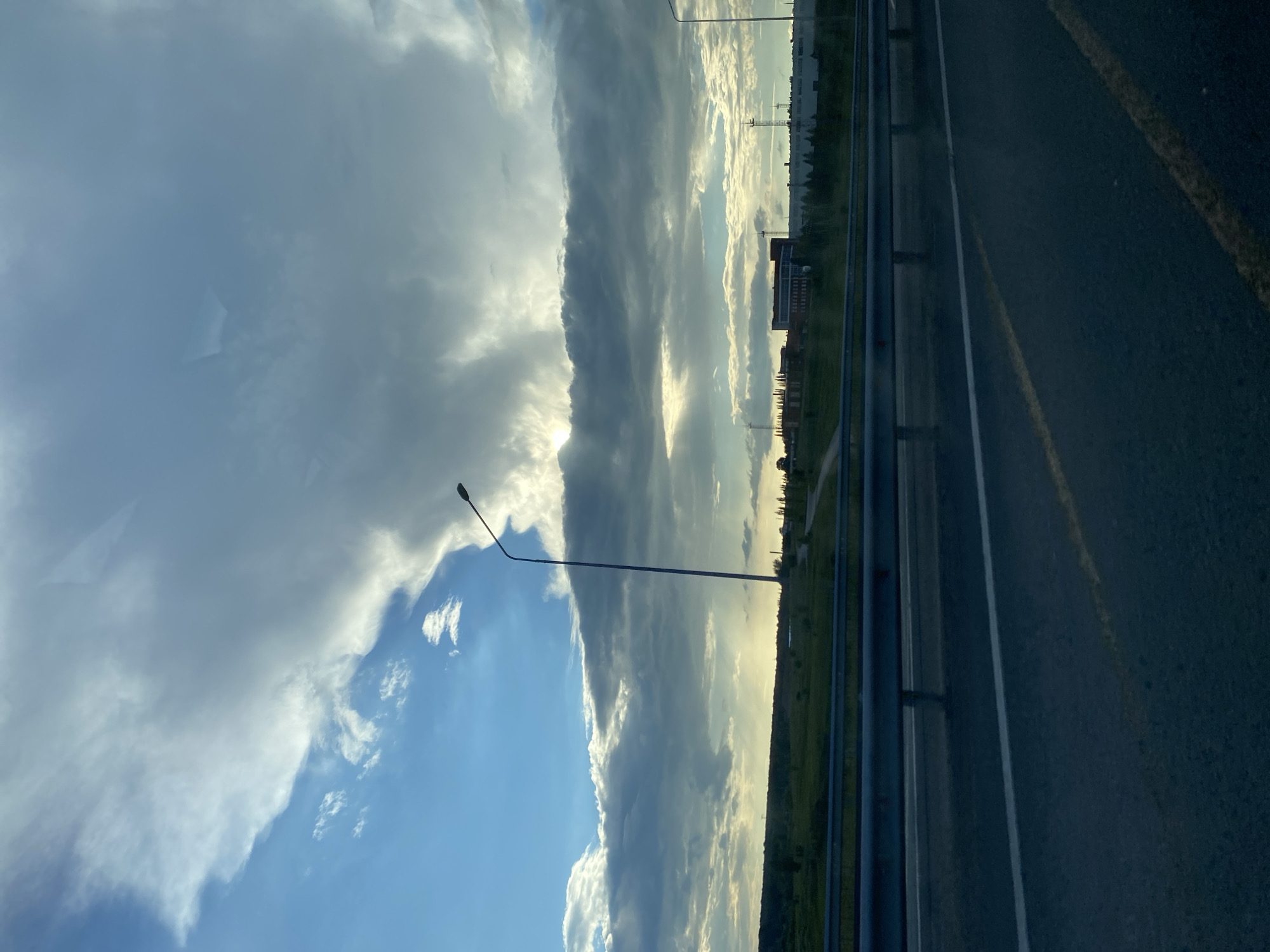 A frontal view from a vehicle moving on an asphalt road shows a vast expanse of sky filled with dramatic, dark gray clouds and patches of bright blue. Sunlight pierces through the clouds in some areas, creating rays of light. In the distance, a flat landscape with green fields and trees is visible. A tall, slender street lamp stands on the left side of the road, a metal guardrail separates the road from the field, and several buildings, including a multi-story brick structure and industrial facilities with towers, are scattered across the horizon. The road itself has white and yellow lane markings. The overall impression is a cloudy, possibly late afternoon or early evening scene on the outskirts of Solov'evka, Russia.