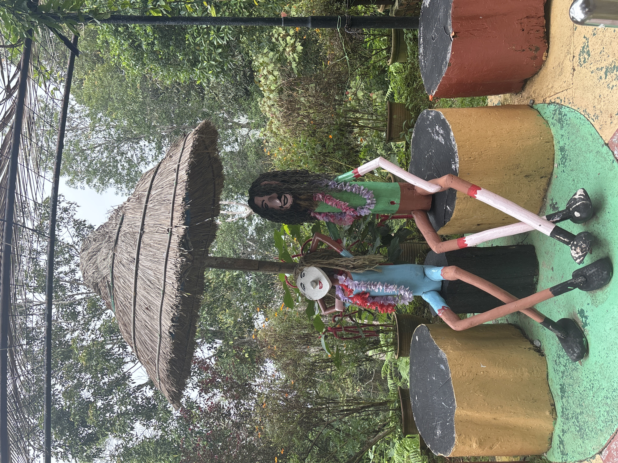 This image depicts a whimsical outdoor scene in George Town, Malaysia, likely a park or garden area. Two colorful, life-sized sculptures of people are seated on large, cylindrical, painted stumps that resemble tree stumps. The sculpture on the left is dressed in a blue outfit and has a round face with drawn-on features. It wears a lei made of red and white flowers. The sculpture on the right is dressed in green and has long, dark, stringy hair and a beard. It also wears a lei, this one with pink and white flowers. Both figures have long, thin, jointed limbs and are posed with their legs crossed.
Above the figures, a large, thatched umbrella provides shade. The umbrella is supported by a thick wooden pole. The background is filled with lush greenery, including trees, shrubs, and potted plants, suggesting a tropical environment. The ground is painted in a playful pattern of green and yellow with some red accents. The overall impression is one of a creatively decorated, outdoor recreational space.