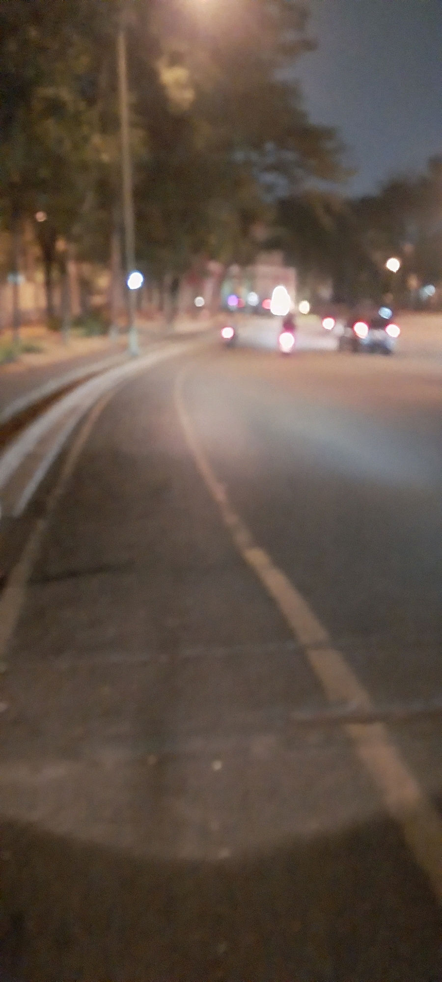 The image shows a blurry, low-light view of a street at night in Kelapa Dua, Indonesia. A curved road is visible in the foreground, with what appears to be tram tracks or a similar parallel structure on the left. A yellow line marks the center of the road. In the background, several out-of-focus vehicles, including what appear to be motorcycles and a car, are present. Trees line the side of the road, and streetlights cast a soft glow, contributing to the overall blurred effect. The scene captures a typical night-time urban environment.