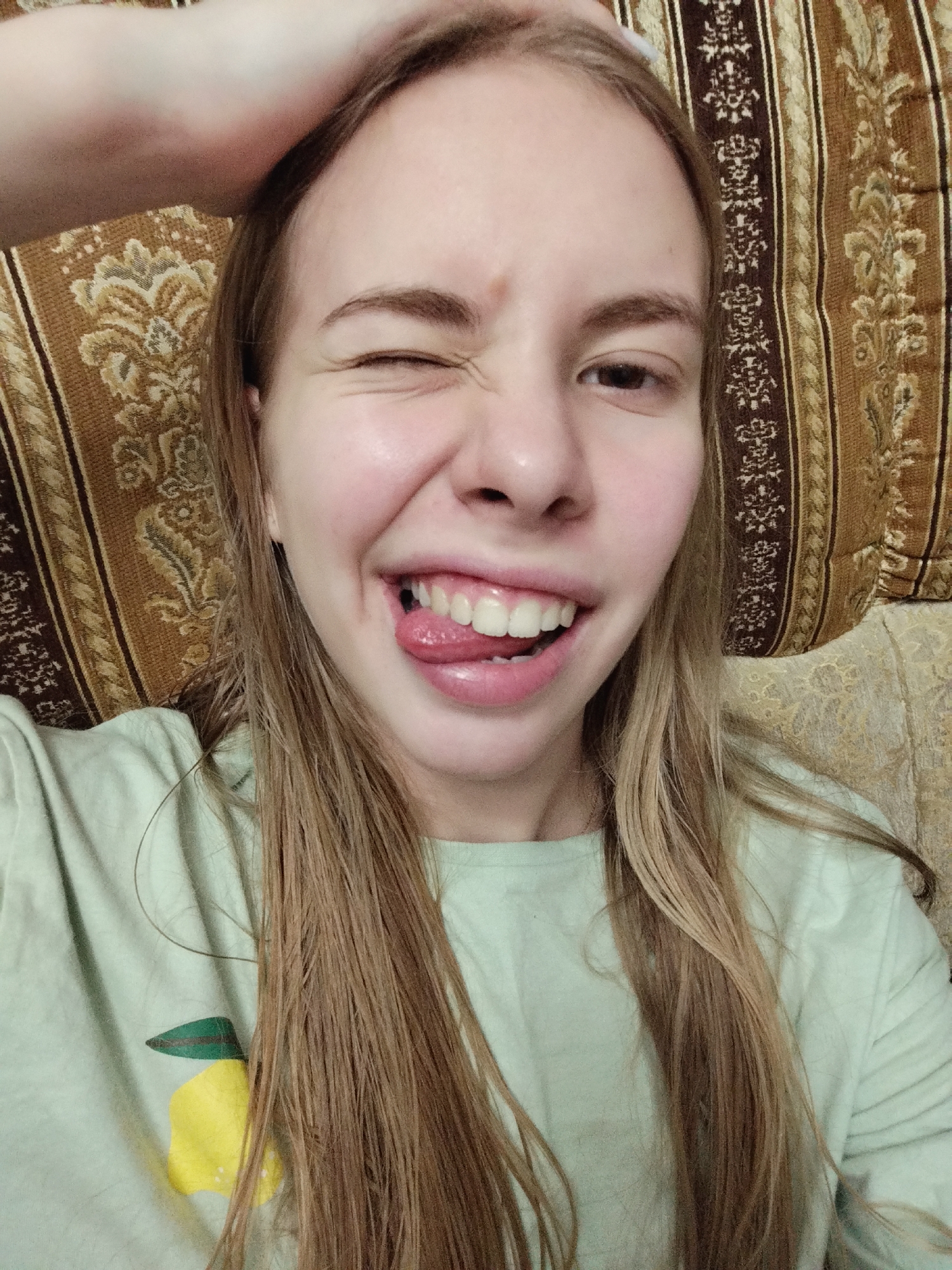 A young woman is taking a selfie. She has blonde hair and is wearing a light green t-shirt with a yellow lemon graphic. Her right eye is closed in a wink, and her tongue is sticking out. Her left hand is raised and resting on top of her head. She is sitting on a couch with a patterned fabric background.