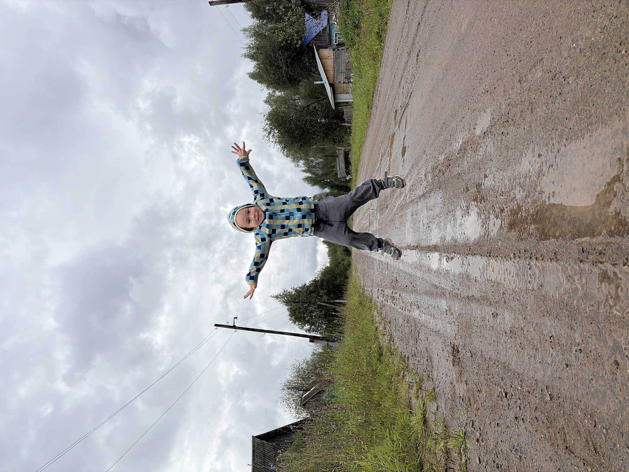A young child in a hooded jacket and grey sweatpants is captured mid-air, jumping on a wet, muddy dirt road. The child's arms are outstretched, and their legs are bent as if in mid-leap. The road is lined with sparse vegetation and appears to be in a rural setting. In the background, there are several wooden houses and trees, characteristic of a village. Power lines are visible stretching across the cloudy sky. The scene suggests a casual, outdoor activity in a somewhat rustic environment. The location is identified as Узяр, Russia.