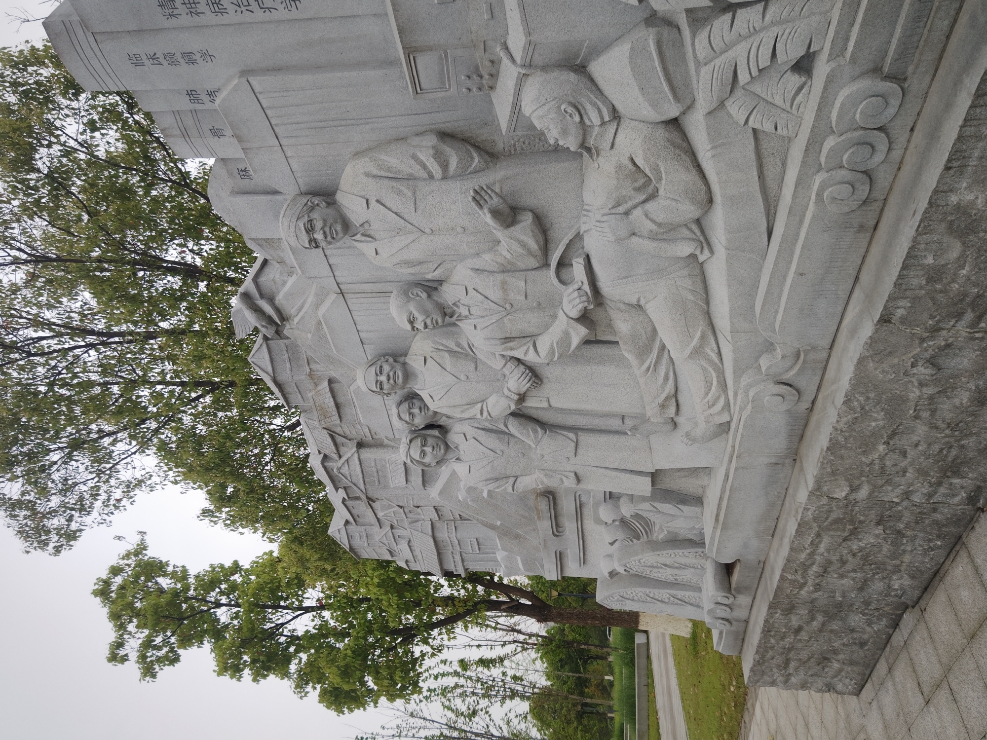 The image shows a stone carving of several figures in Chongqing High-tech Zone, China. The figures appear to be doctors or medical personnel, depicted in various poses, possibly representing aspects of medical practice or history. The carving is positioned outdoors, with trees visible in the background. In the background some chinese letters are visible. The carving has a rough, textured base. The setting appears to be a park or public area within the high-tech zone. The sky is overcast. A pathway is visible.