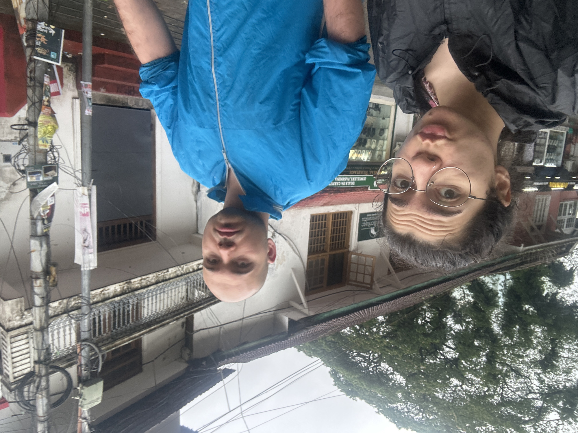 This image features two men against an urban backdrop, likely in Kochi, India. The photo is taken from a slightly low angle, showing them looking towards the camera.
The man on the left has a shaved head and wears a bright blue waterproof jacket with a white zipper. The man on the right wears a black jacket and round, metal-rimmed glasses, with dark, shoulder-length hair. Both individuals have neutral expressions, looking directly at the camera; the man with glasses has slightly furrowed brows.
Behind them, the setting includes buildings with traditional balconies and wooden-framed windows. To the left, a utility pole is heavily laden with a dense network of wires and various posters, some faded and illegible. One white sign with green lettering on a building clearly displays parts of "JEWELLERY, FASHION" and a logo that appears to say "Heritage". Other smaller signs on the utility pole are visible, one possibly reading "NP".
The sky is overcast, indicating a cloudy daytime setting without direct sunlight. Large, green trees are visible against the sky on the right side of the frame. The jackets suggest either cool weather or protection from potential rain. The architecture and street elements are consistent with an urban environment in South Asia.