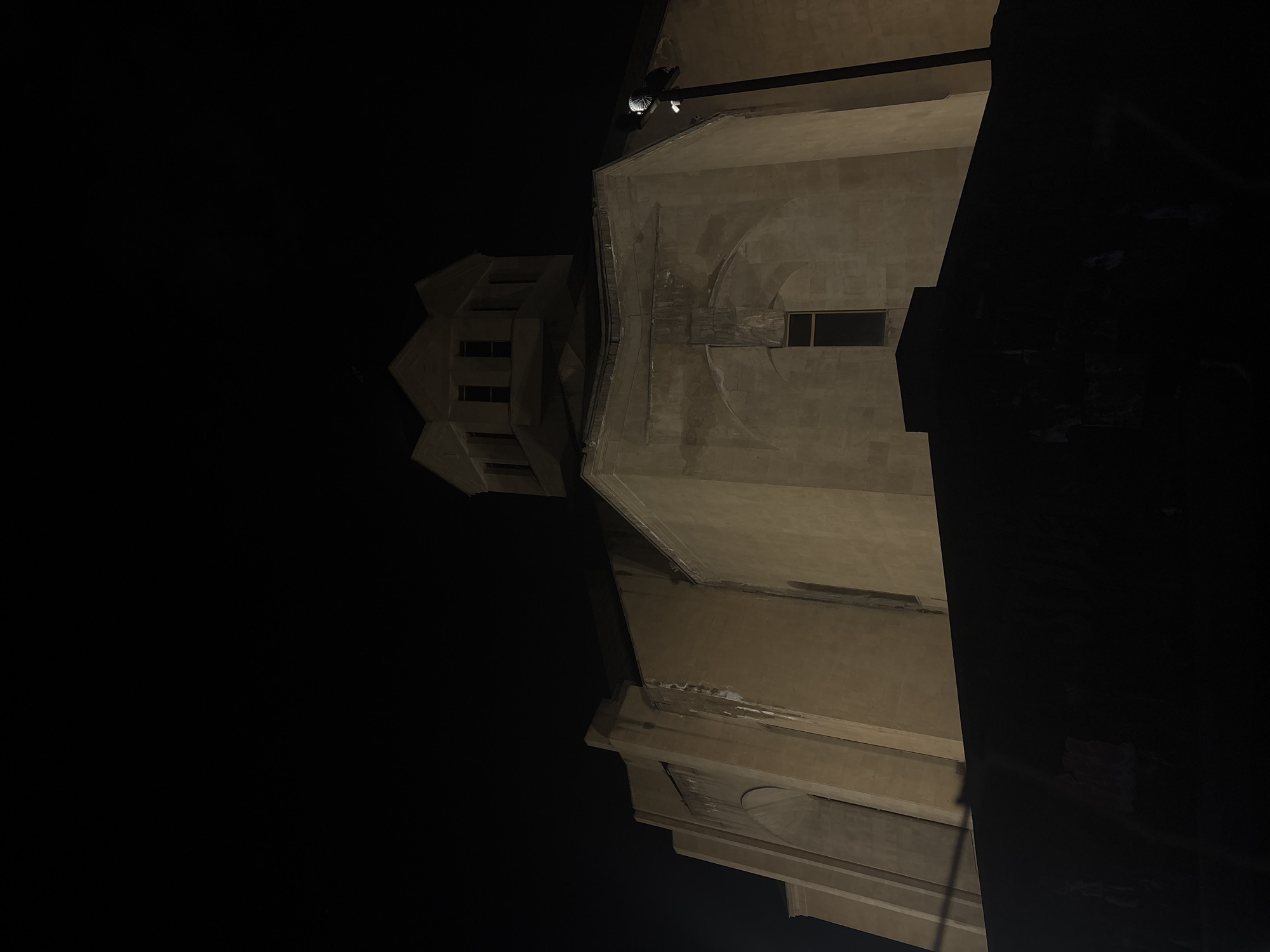 The image shows a low-angle view of a building at night. The building appears to be constructed of stone or concrete, with architectural features including windows and what looks like a tower or spire. The lighting is dim, with most of the scene in shadow, indicating that it is nighttime. Given the information from the prompt, the image is likely of a building located in the city of Yerevan, Armenia. There are no people visible in the image. The focus is on the architectural details of the building illuminated against the night sky.