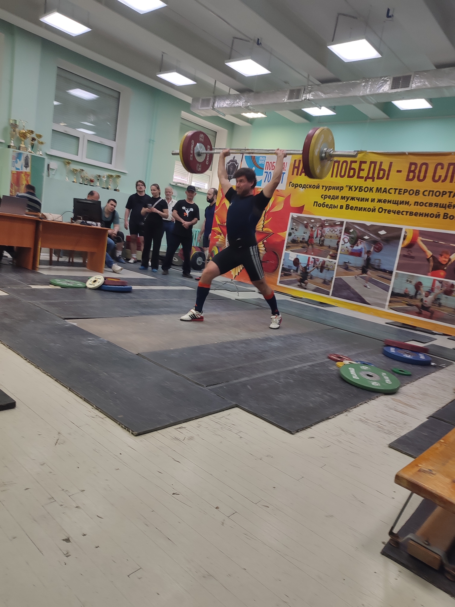 Here is a description of the image:
A weightlifter is in the middle of a lift, seemingly completing a clean and jerk, with the barbell overhead. He is positioned on a weightlifting platform in what appears to be a gymnasium or sports hall. A small group of spectators stands nearby observing the event. Behind the lifter, there is a banner with Russian text and images of other weightlifters. The banner seems to celebrate the "Victory in the Great Patriotic War" and mentions a tournament called "Masters of Sports Cup." The gymnasium has a window with trophies on display near a table with a computer and people sitting at it. Based on the text and context, the location is in Izhevsk, Russia, as identified by the Russian text on the banner.