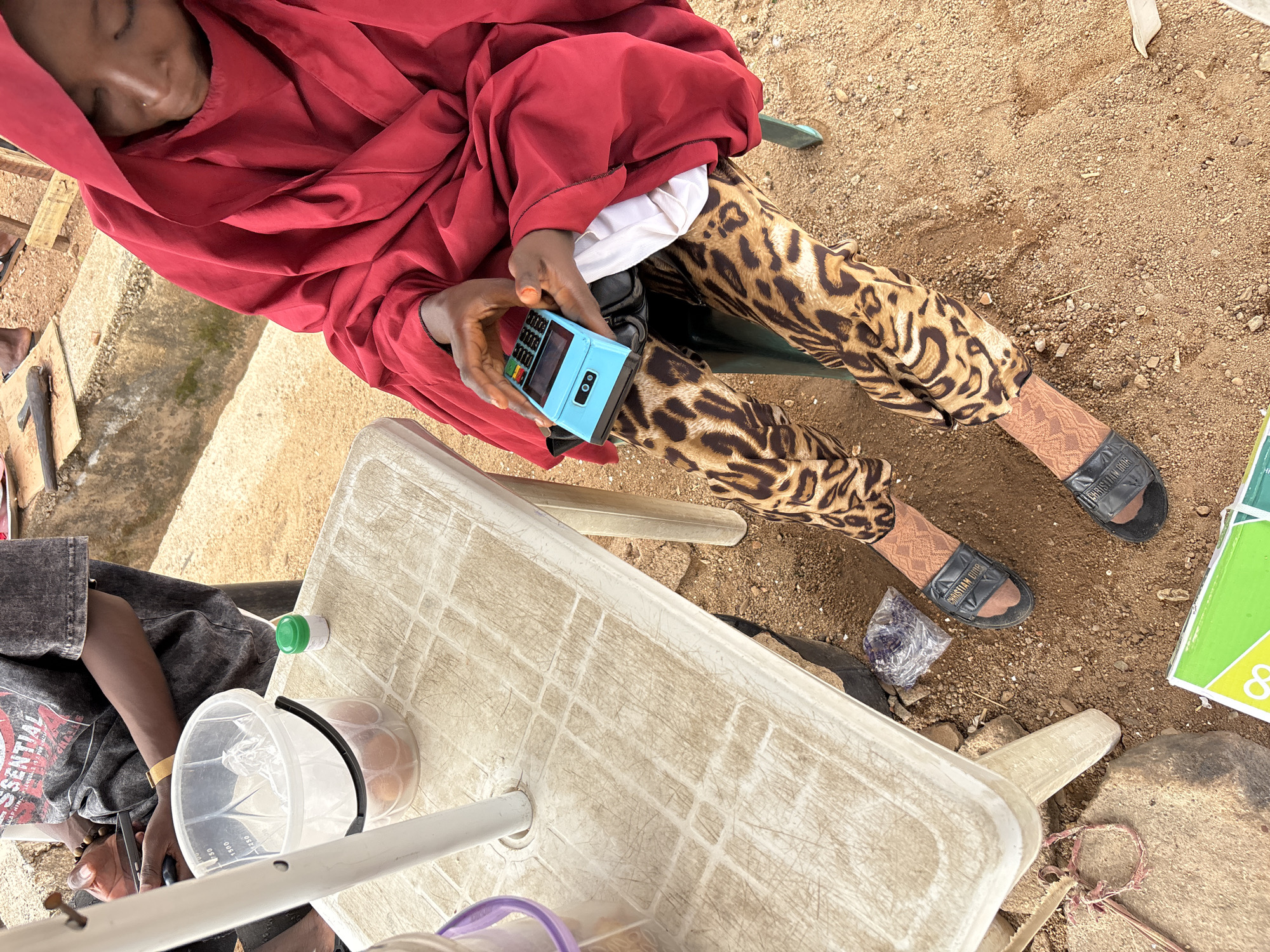 The image shows a person, appearing to be a child or young person, wearing a red hijab and leopard print trousers. They are sitting outdoors on a dirt ground, with a small, light blue electronic device, possibly a card reader or payment terminal, held in their hands. The device has a keypad and a small screen. The person is looking down, seemingly engaged with the device.
Beside the person, to their right, is a light-colored, plastic table. On the table, there are a few items, including a white plastic bucket with a black rim containing some round objects, possibly eggs or fruit. A small white bottle with a green cap is also on the table.
To the left of the person, part of their body is visible, wearing a dark grey t-shirt with white text that reads "ESSENTIAL". The background consists of sandy, uneven ground with some small rocks and debris, suggesting an outdoor setting in a possibly rural or informal urban environment. The lighting suggests it is daytime. There are no clear indications of the weather, but the ground appears dry. The overall impression is a candid shot of a transaction or interaction involving the electronic device.