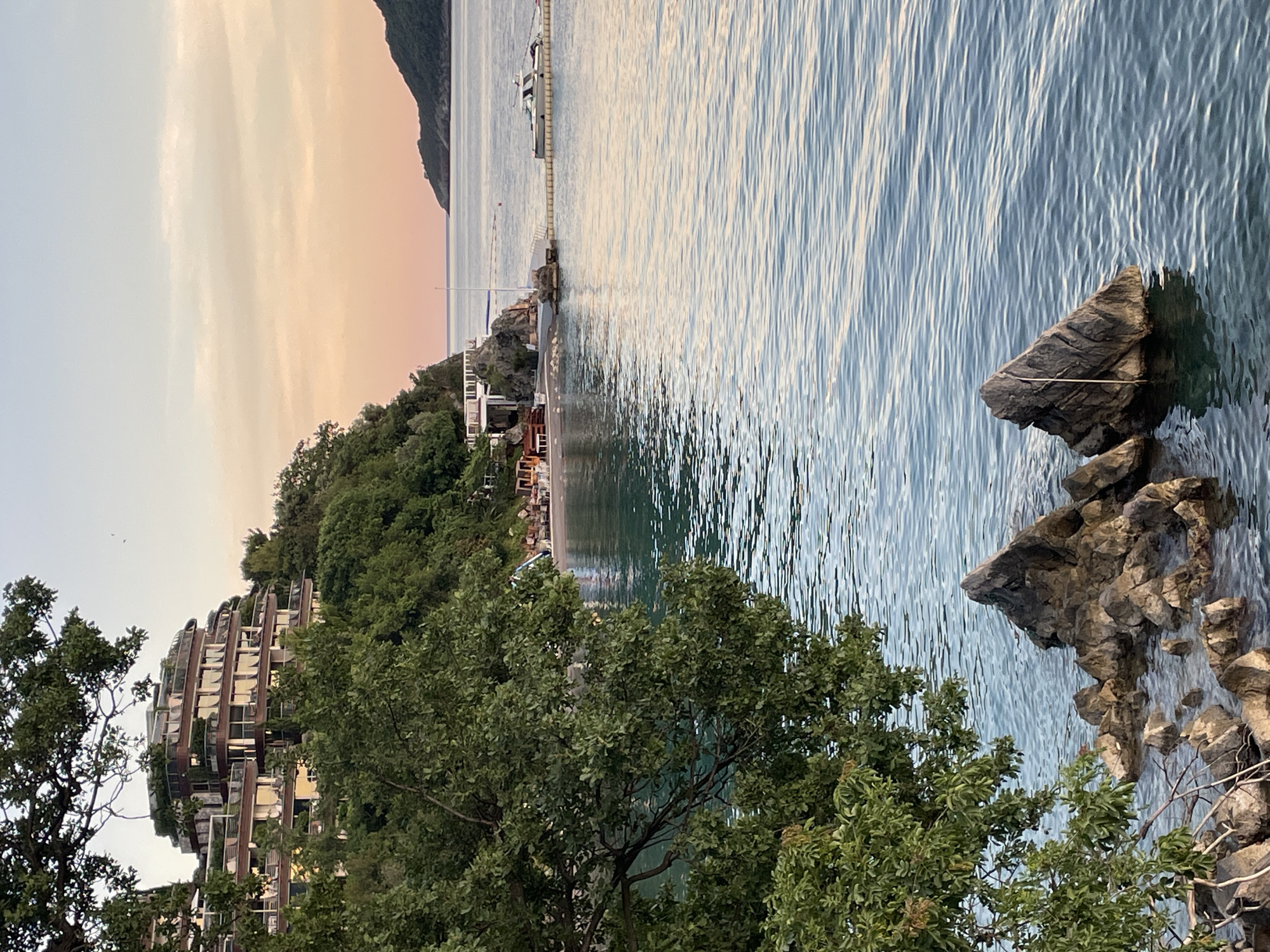 The image depicts a scenic coastal view in Budva, Montenegro. In the foreground, a cluster of jagged rocks emerges from the calm, turquoise water, with a large, leafy tree branch extending from the left side of the frame, partially obscuring the view.
In the midground, a small, pebble beach is visible, with a few scattered tables and chairs set up, suggesting a cafe or restaurant. A white structure with balconies and a railing is built on a rocky outcrop overlooking the beach. A small white boat is moored to a yellow floating barrier in the water near this structure.
In the background, a multi-tiered building with a distinctive curved, layered design is nestled on a lush green hillside. This building features numerous windows and balconies, with hints of warm interior lighting. The overall scene is set against a soft, pastel-colored sky, with hues of pink and orange blending into a pale blue, indicating either sunrise or sunset. In the far distance, a dark, tree-covered island or peninsula rises from the sea. The water is serene, with gentle ripples reflecting the sky and surrounding landscape.