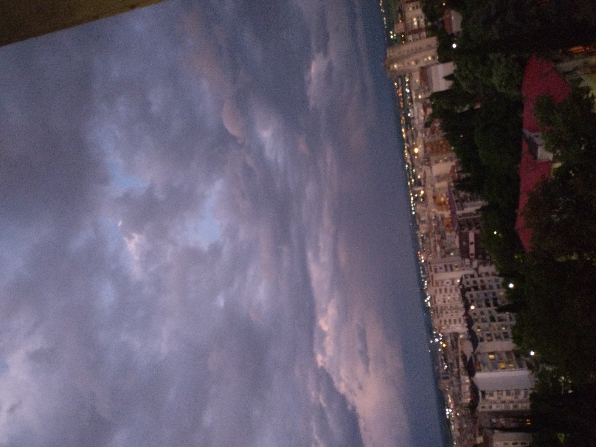 The image shows a nighttime view of Stara Zagora, Bulgaria. The sky is filled with dark, ominous clouds. In the distance, a city skyline is visible with numerous buildings illuminated with lights. Near the city, there is a dark line of trees. The image appears to have been taken from an elevated position.