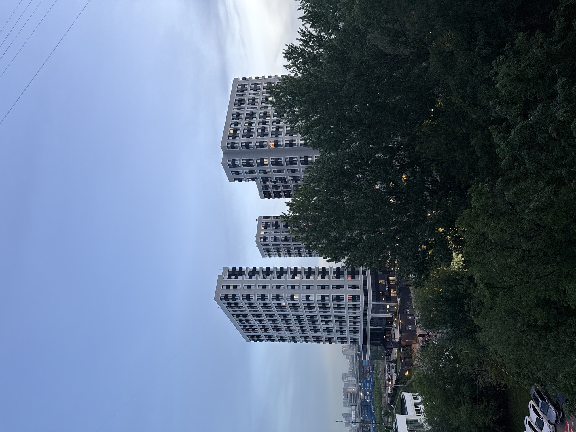 The image shows three modern apartment buildings in Khimki, Russia. The buildings are multi-story and feature a grid-like pattern of windows and balconies. Lush green trees partially obscure the view of the buildings and the surrounding cityscape. In the distance, a hazy skyline indicates the presence of other buildings. Overhead, power lines are visible against a pale blue sky. The scene appears to be taken at dusk or dawn, as the light is soft and diffused.