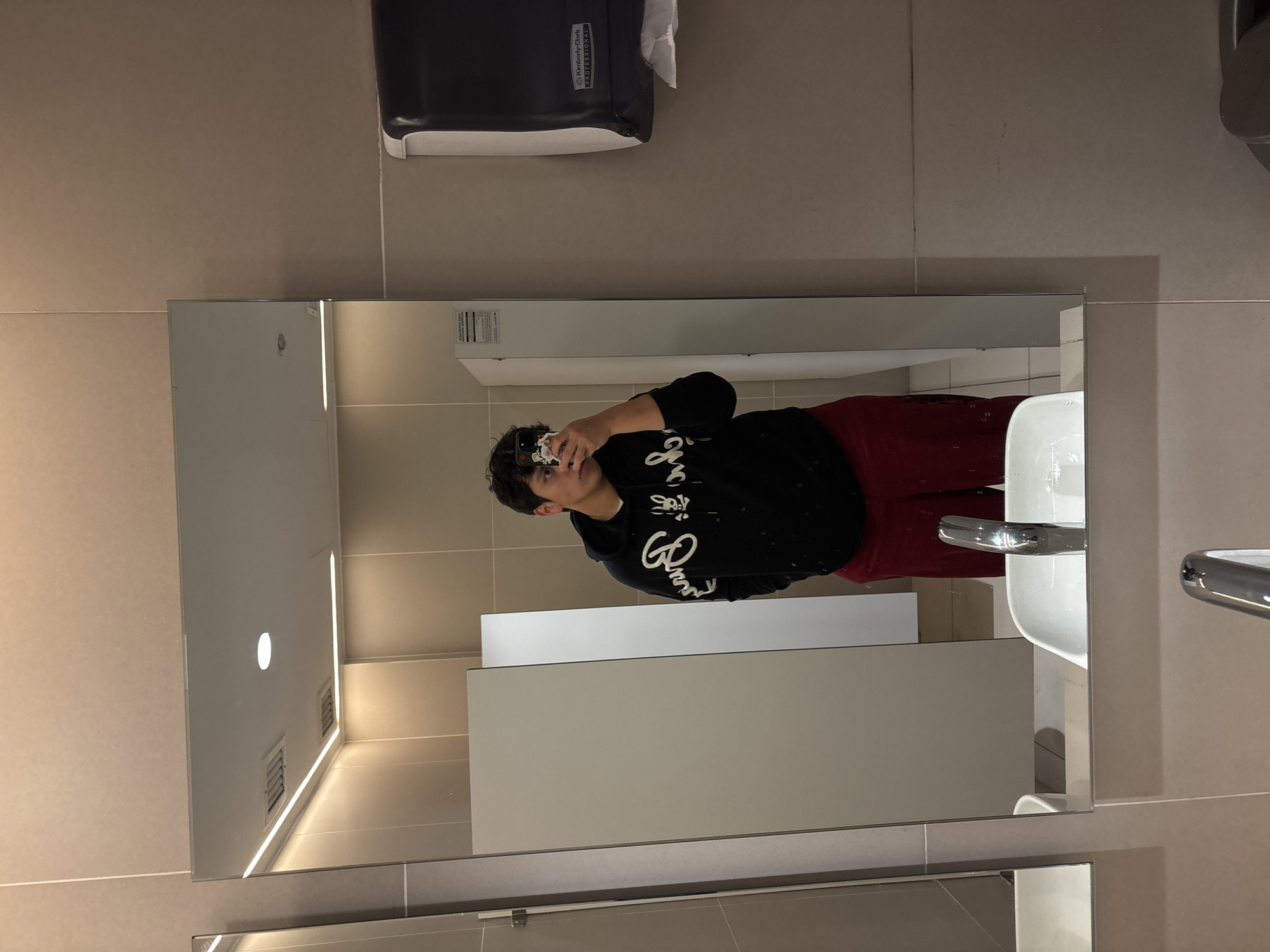 A person wearing a black hoodie and red pants is taking a picture of themselves in a bathroom mirror. The bathroom has light brown tiles, a black paper towel dispenser, a white sink, and restroom stalls. The location appears to be somewhere in Mexico City, Mexico.