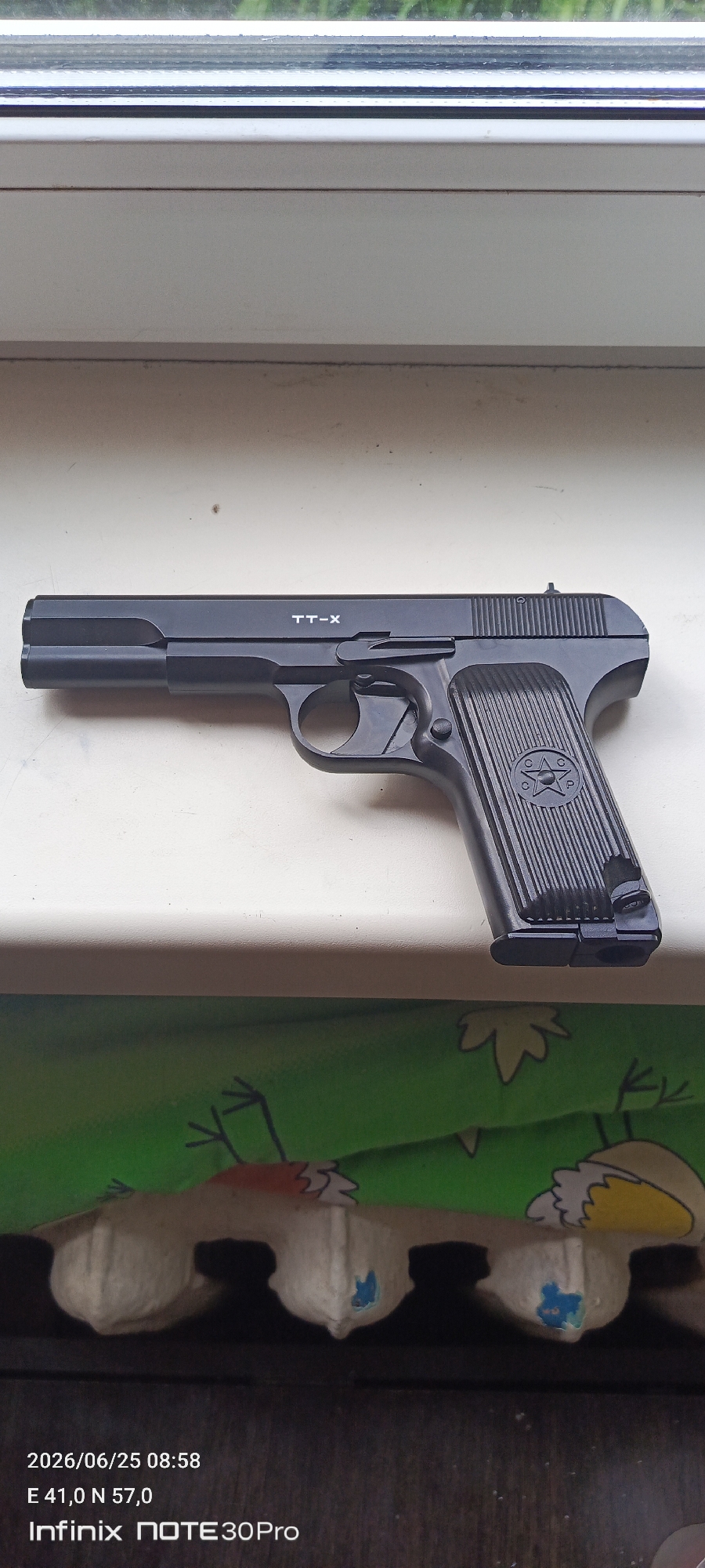 A black handgun is placed on a white windowsill. The handgun is a TT-X model. It has a ridged grip with a star emblem. The scene is indoors, as suggested by the windowsill and the presence of a window behind it, which shows green foliage and a blurred sky. Below the windowsill, a colorful, cartoon-like pattern is visible on fabric, and what appears to be part of an egg carton. The lower portion of the image displays text overlay, indicating the date "2026/06/25 08:58", coordinates "E 41,0 N 57,0", and "Infinix NOTE 30Pro". The time and the ambient lighting suggest it is daytime. No people are visible in the image. The overall impression is a still-life arrangement, possibly a product photograph or a personal item displayed. The location context provided is Ivanovo, Russia, which aligns with the coordinates and potential origin of the text on the screen.