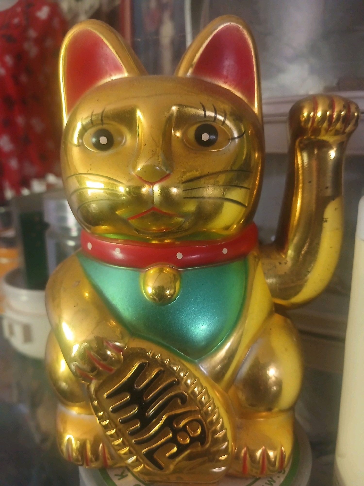 A close-up, eye-level, medium shot captures a golden, beckoning cat figurine. The cat, a Maneki-neko, is positioned centrally in the frame, facing slightly to the left. Its left paw is raised in a welcoming gesture. The figurine is a lustrous gold color, with red inner ears and accents on its collar. A green bib adorns its chest, featuring a small, gold-colored charm. The cat holds a dark, ornate object, possibly a coin or fish, in its front paws. The background is blurred, with hints of red fabric and other indistinct objects, suggesting an indoor setting, possibly a shelf or display. The lighting is warm, with noticeable reflections on the metallic surface of the figurine, indicating a glossy finish. There are no people visible in the image, and no actions or interactions are taking place. The scene is static and focuses solely on the decorative cat figurine. The provided location information, "Banyuwangi, Indonesia," is not visually represented in the image itself.