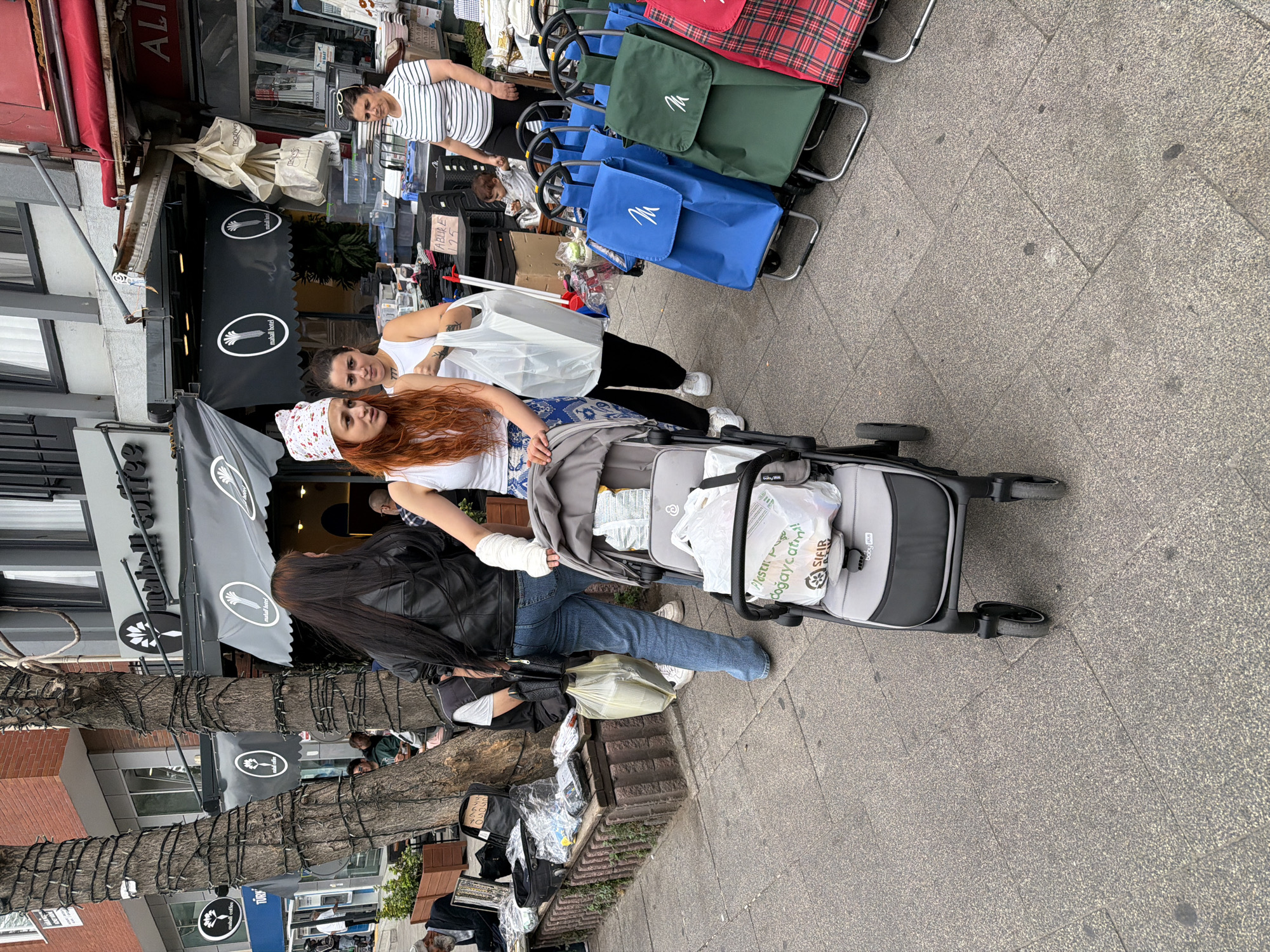 The image captures a street scene in Kadıköy, Turkey, likely near a shopping area given the presence of various vendors and pedestrians. In the foreground, a woman with long red hair, wearing a white tank top and patterned pants, pushes a gray baby stroller. Her left arm is bandaged. Beside her, another woman in a white tank top carries a large white shopping bag. Behind them, a woman with long dark hair, wearing a black leather jacket and blue jeans, walks away from the camera.
The background reveals a street lined with shops, one of which has awnings and displays goods. Multiple shopping trolleys, some blue and green, are visible near the shops, suggesting a marketplace or an area with portable vendors. A baby is seated in a shopping cart in the middle ground. Several trees are present, decorated with what appear to be string lights. The ground is paved with stone tiles, and there are some potted plants and benches along the sidewalk, where various items are laid out for sale. The overall atmosphere suggests a bustling urban environment with commercial activity.