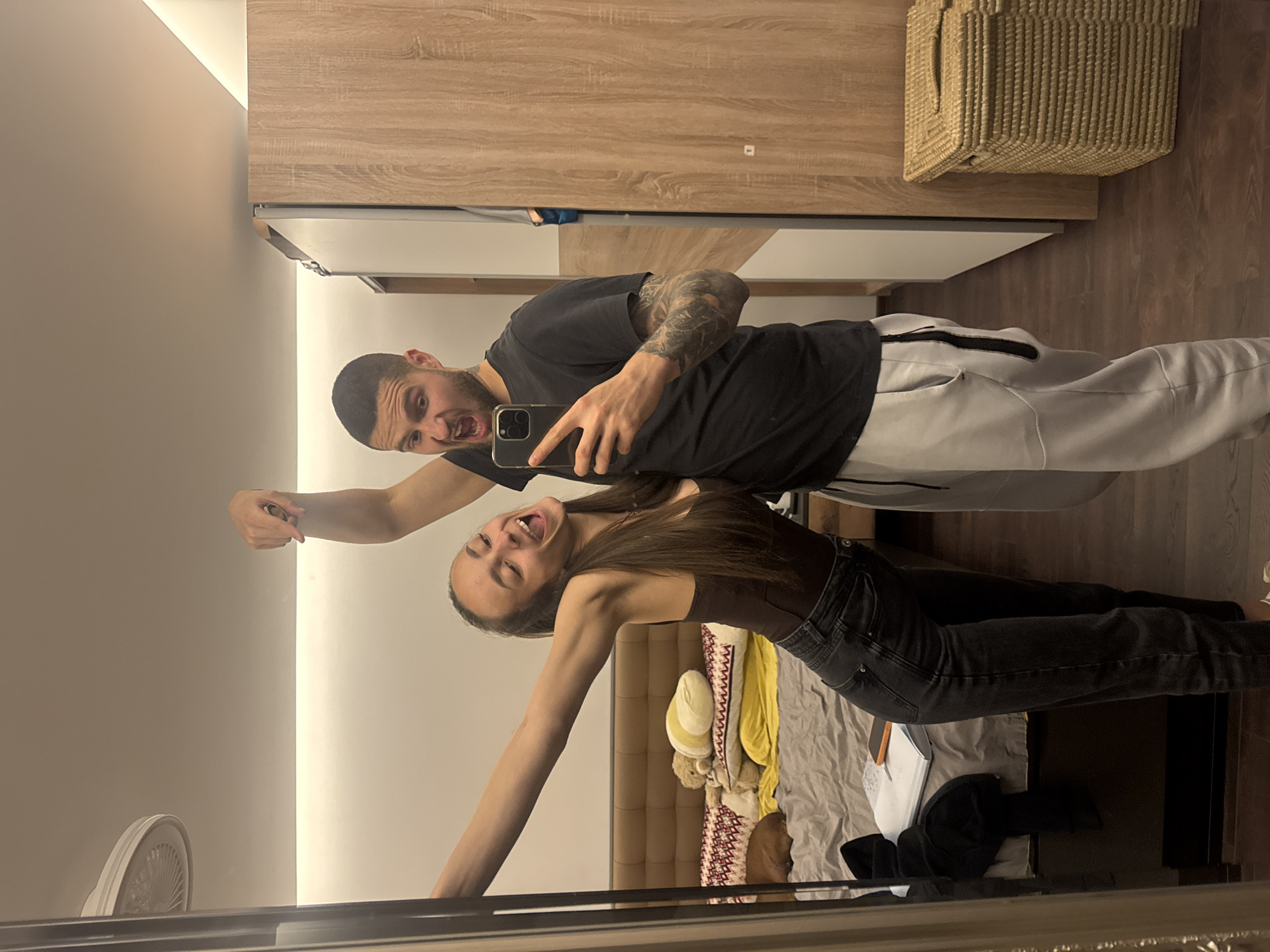 A selfie taken in a mirror shows a man and a woman. The man is on the left, wearing a black t-shirt and gray sweatpants, holding a phone in his right hand. He has short dark hair and a tattoo on his left arm. The woman is on the right, wearing a brown top and black jeans. She has long brown hair and is sticking her tongue out. They appear to be in a bedroom. The room includes a bed with various items on it, a wooden wardrobe, and a wicker basket on top of the wardrobe. The presence of a European style electrical outlet on the wall suggests the location is in Europe. Based on the user's prompt, the city is identified as Sofia, Bulgaria.