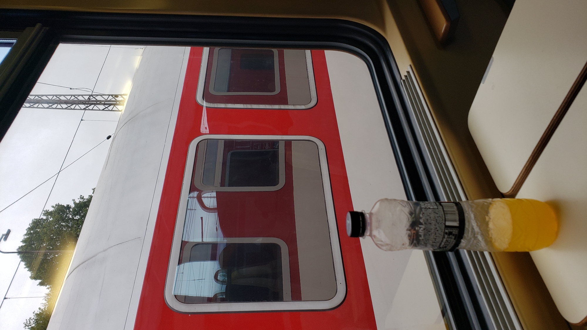 A slightly angled, medium shot captures a view from inside a train carriage, looking out a window at another train. In the foreground, a plastic bottle filled with a yellow liquid, likely juice, stands on a folded-down tray table. The bottle has a black cap and a label with text and images. The tray table is light beige with a wooden trim. The window frame is dark, and the outside is dominated by the exterior of a red and white train. The red section has several rectangular windows, some of which reflect the interior of the train. Beyond the train, an overcast sky is visible, along with trees and a metallic pylon with overhead wires. The lighting is diffused, suggesting an outdoor scene on a cloudy day. The composition creates a sense of travel and contemplation. The location is the train station in Vrútky, Slovakia.