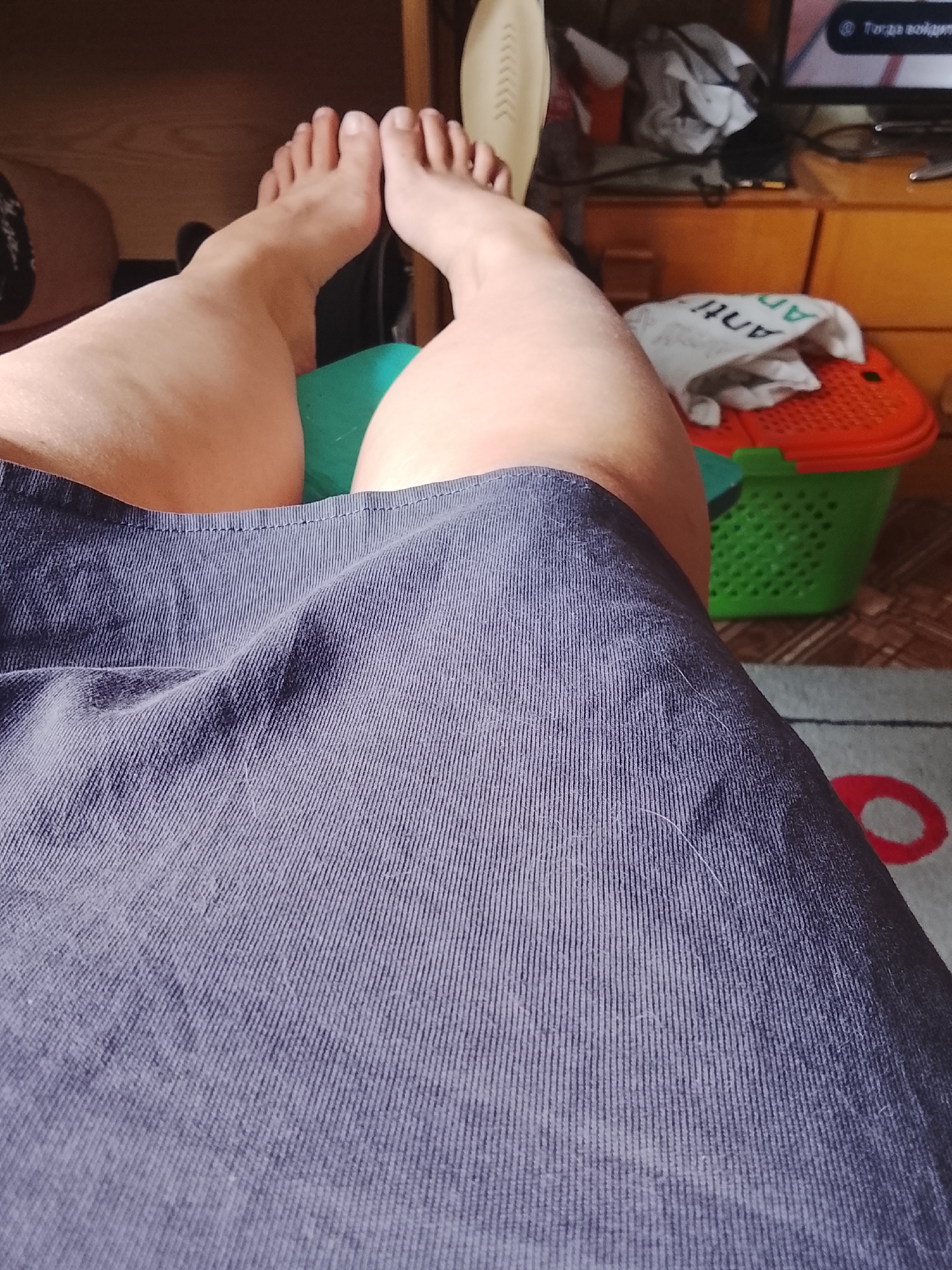 The image shows a close-up view of a person's legs, from the knees down, covered by a dark blue, ribbed fabric, possibly a skirt or dress. The feet are bare and visible, with toes pointing upwards. In the background, partially visible objects include a green and orange plastic basket, a television screen, and wooden furniture. The scene appears to be indoors, in a domestic setting. There is no specific indication of the location within Almaty, Kazakhstan.