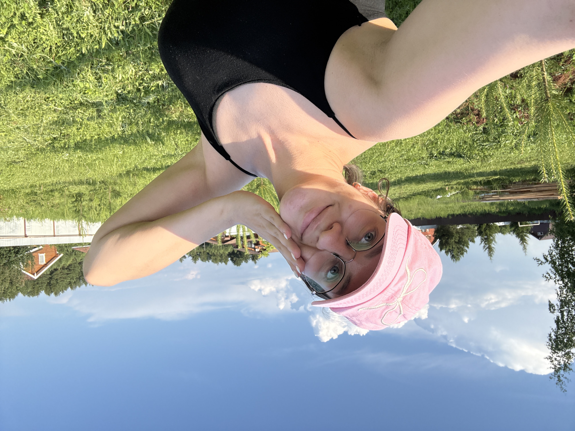 A woman wearing a pink baseball cap and black tank top takes a selfie outdoors in a grassy area. She is smiling and touching her face with her hand. In the background, there are trees, a fence, and a few houses, suggesting a rural or suburban setting. The sky is blue with white clouds.
