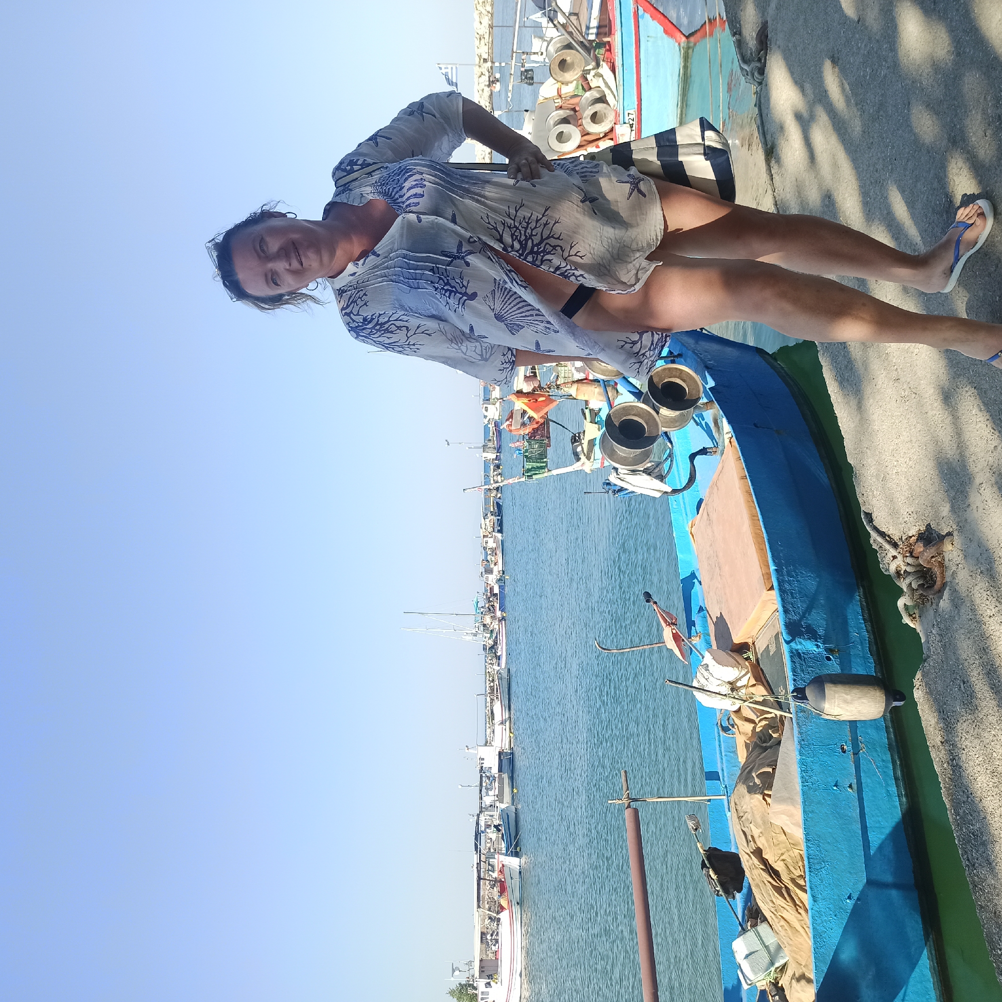 A medium shot captures a woman standing on a concrete dock in the harbor of Limani Litochorou, Greece. The woman is positioned to the right of the frame, facing towards the viewer with her right hand on her hip. She is wearing a white, sheer cover-up with a blue coral-like pattern, which is open to reveal dark swimwear underneath. She has sunglasses perched on her head and her hair is pulled up. Her legs are bare, and she is wearing blue and white flip-flops. To the left of the woman is a blue fishing boat with various pieces of equipment, including nets and buoys, on board. The boat is partially moored to the dock. In the background, several other fishing boats and sailboats are visible in the calm blue water of the harbor. A clear blue sky with no clouds forms the upper part of the image. The sunlight casts distinct shadows on the concrete dock from the woman and the boats. The overall scene depicts a sunny day at a Greek harbor with a woman posing by a fishing boat.