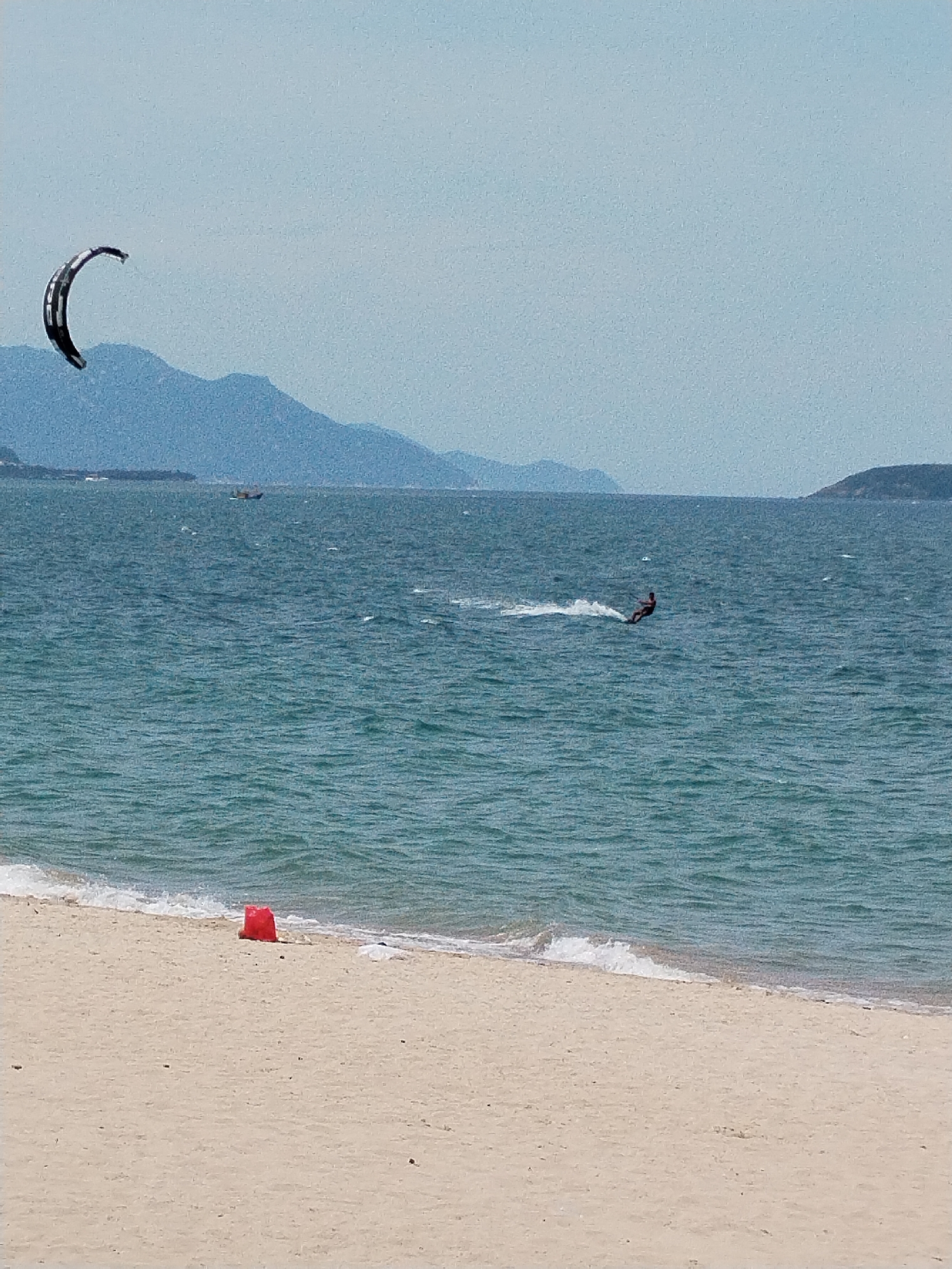 The image depicts a bright, sunny day at a beach in Nha Trang Ward, Vietnam. The foreground is dominated by a sandy beach with a few dark specks scattered across it. A small, bright red object, possibly a bucket, rests near the waterline. Gentle waves lap the shore, creating a white foamy edge.
In the mid-ground, the turquoise sea stretches out, with visible ripples and waves indicating a moderate breeze. A lone kitesurfer is actively engaged in the sport, carving a white trail of spray as they ride across the water. In the distance, a small boat is visible on the horizon, and beyond that, a range of hazy blue mountains forms the backdrop, with some landmasses or islands visible on the right. The sky is a pale blue, clear except for a slightly hazy quality, suggesting the presence of a distant haze or light cloud cover. A large, dark kitesurfing kite is visible in the upper left quadrant of the image, suspended against the sky and the distant mountains.