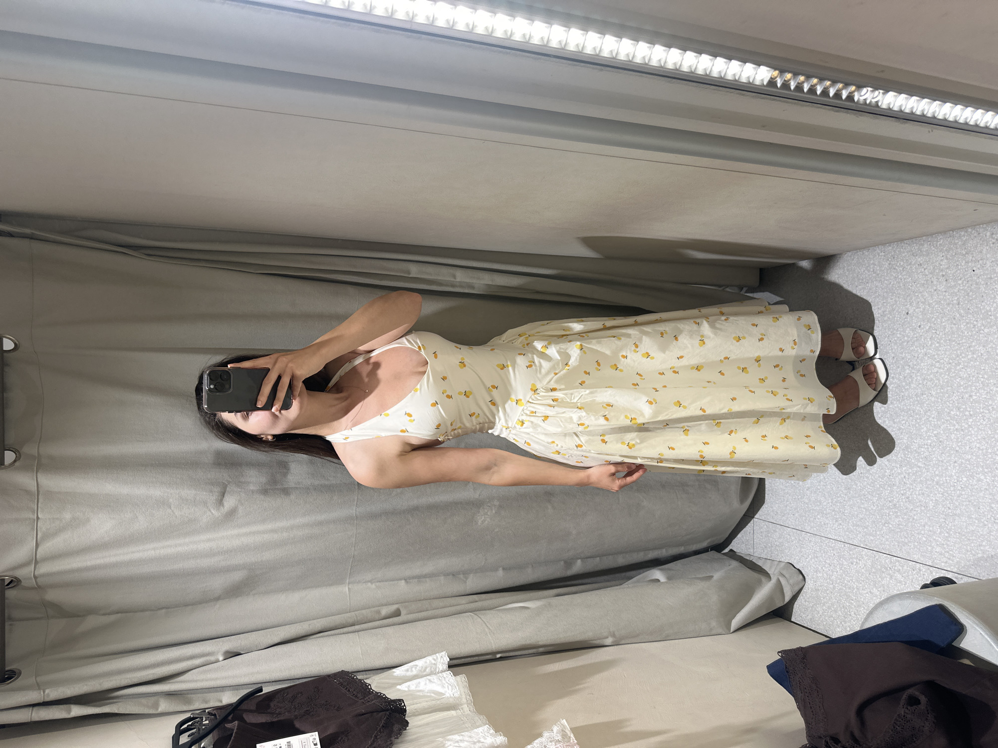 A woman is taking a mirror selfie in a changing room. The room is lit by a fluorescent light panel on the ceiling. The woman is wearing a light-colored floral print dress and white sandals. The changing room has a gray curtain and speckled gray floor tiles. On the bench in the changing room, there are items of clothing in dark colors, including a brown piece of clothing with lace detail.