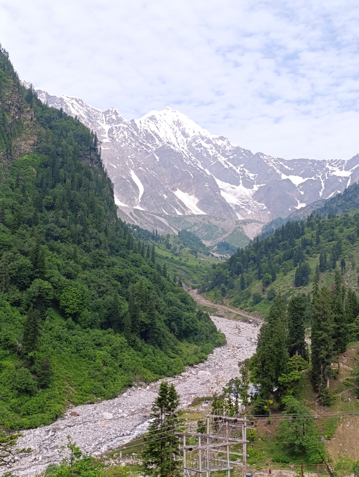 The image shows a majestic mountain landscape with a rocky riverbed in the foreground. Dense green forests cover the slopes of the mountains, transitioning to snow-capped peaks in the distance. The sky is partly cloudy, with patches of blue visible. There are no people or animals visible in the image. In the bottom right corner, there is an electrical substation with power lines. The scene appears to be a natural vista, possibly in a mountainous region like Dhundi, India, known for its scenic beauty.