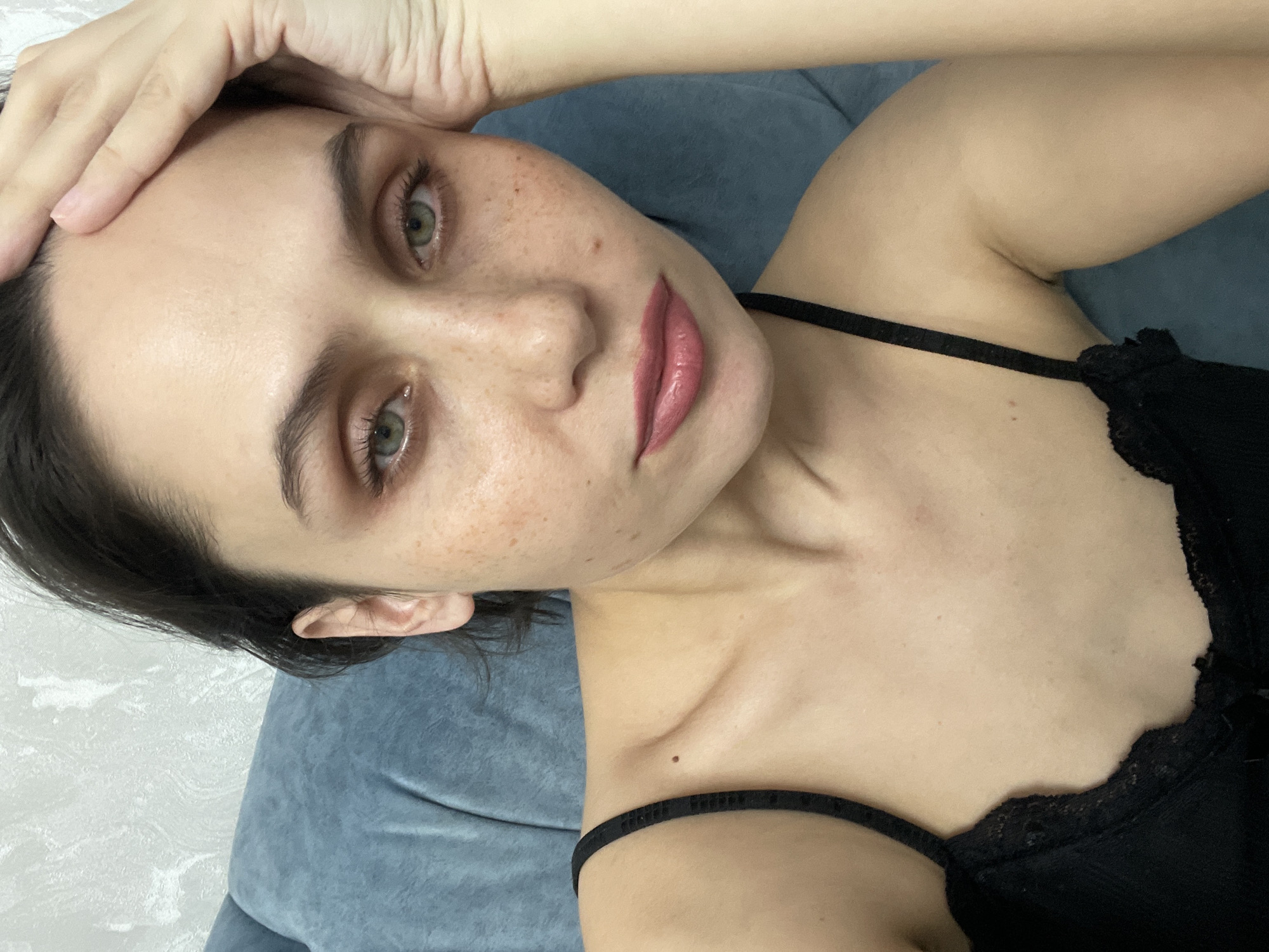 An indoor portrait features a young woman lying on her back on a blue sofa, looking directly at the camera. Her head is angled towards the left, with her left hand resting on her forehead. She has dark hair, green eyes, and distinct dark eyebrows. Her skin is fair with numerous freckles visible across her nose and cheeks. Her full lips are tinted with glossy pink lipstick. She is wearing a black top or bralette with thin straps and intricate lace detailing. The background behind her head is a light-colored, textured wall.
The cropped images provide closer views of specific details: her eyes and forehead, her lower face with prominent lips and freckles, her upper arm and shoulder, her ear and dark hair, her collarbone area showing a small mole, and the lace trim of her black top.
The lighting appears to be soft and even, typical of an indoor environment. Her expression is neutral and calm. There is no visible text in any of the images. The scene is set indoors in Izhevsk, Russia.