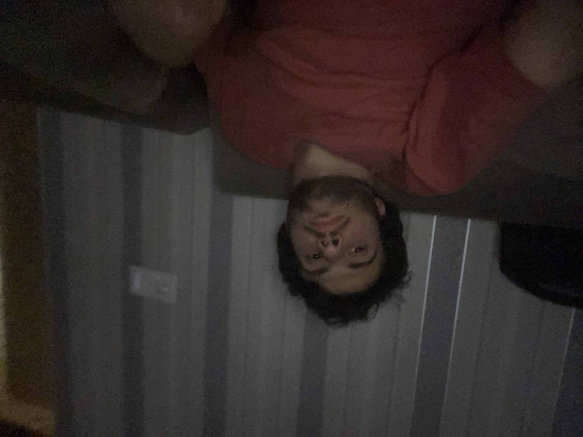 The image shows a close-up, low-angle, self-taken photograph of a young man with dark, curly hair and a beard. He is wearing a red t-shirt and is seated on a couch. Behind him, a wall with vertical, ribbed paneling is visible, with a light fixture or electrical outlet on the right side. The lighting is dim, suggesting an indoor environment, possibly at night. The overall impression is of a casual, personal moment captured by the individual. The background does not provide any specific visual cues to identify a location within Tbilisi, Georgia.
