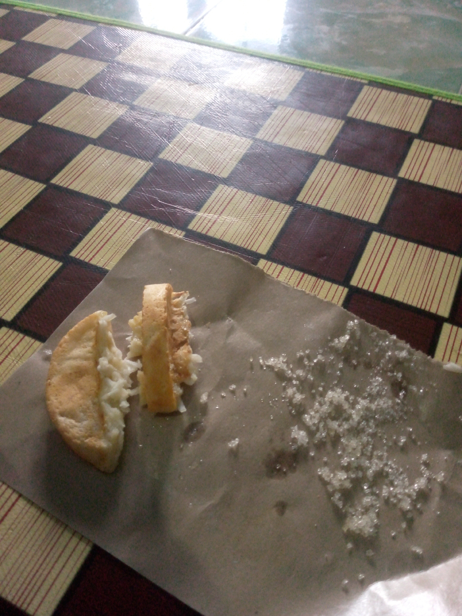 An overhead shot shows a slice of an Indonesian pancake, possibly "kue cubit" or "kue ape," placed on brown paper. The pancake is light brown and appears to have a soft, spongy interior with shredded coconut filling. Next to it, a small pile of white sugar granules is scattered on the paper. The scene is set on a checkered mat, with alternating squares of brown and a light woven material. The mat's pattern suggests a location in Indonesia, possibly Giriasih. The lighting is bright, indicating it's daytime, and the surface appears to be a tabletop.
