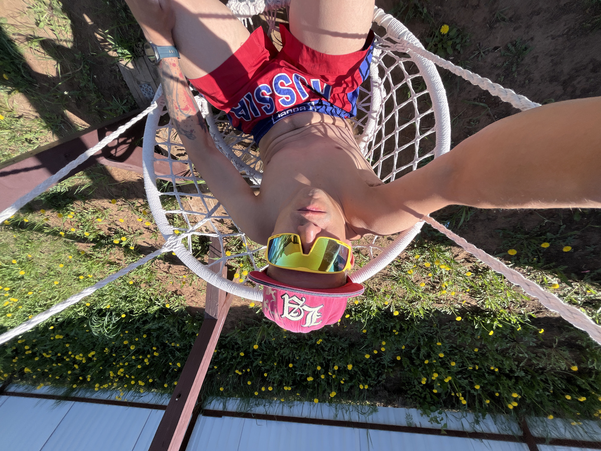 A shirtless individual is lounging in a white, macrame-style chair suspended from a brown wooden structure outdoors. The person is wearing red shorts with "RUSSIA" printed in blue letters and a pink baseball cap with a logo on the front. They are also wearing yellow sunglasses. The scene is shot from a first-person perspective, looking upwards at the individual in the chair. The background consists of green grass dotted with small yellow flowers. In the bottom of the frame, a white metal fence is visible. There is no information given that definitively places the image in the city of Chaikovsky, Russia.
