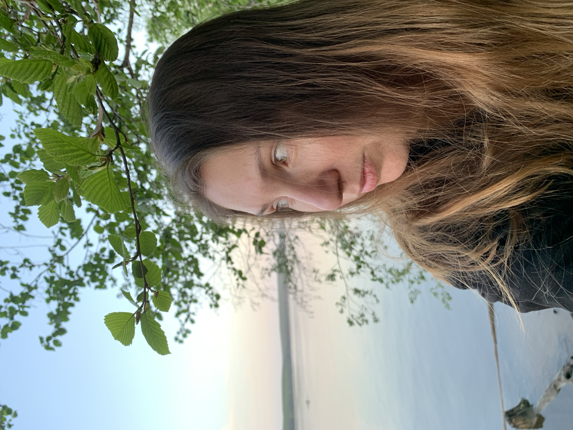 A sideways, low-angle shot of a fair-skinned woman with long brown hair with blonde highlights, who is looking down. A branch with green leaves is to the upper left. In the background is a body of water with a light-colored sky. Based on the location, this could be the city of Chaikovsky, Russia, and the body of water is likely the Kama River.