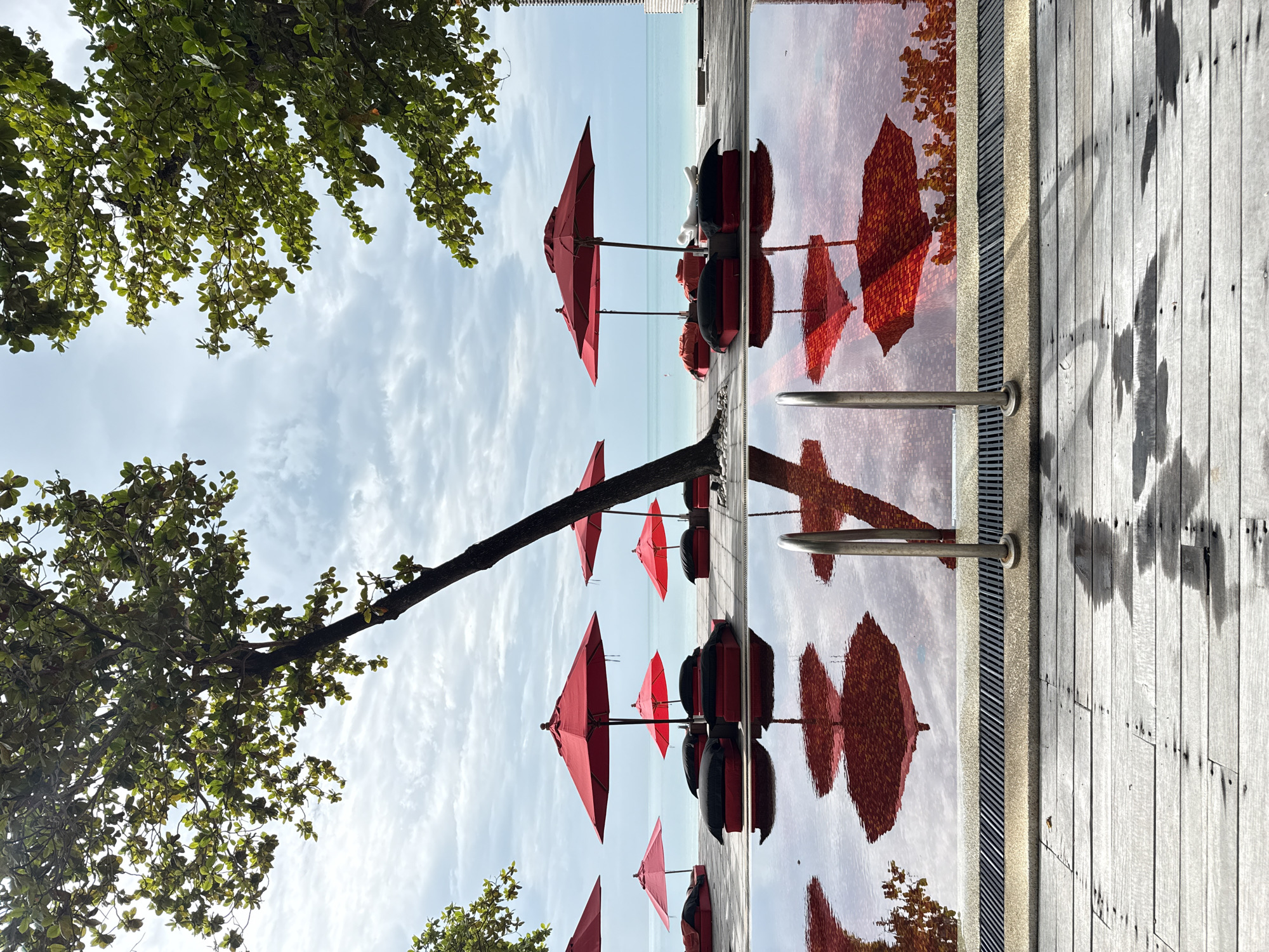 This is a vertical, eye-level, medium-shot photograph of a resort's swimming pool and beach area in Koh Samui District, Thailand. In the foreground, a wooden deck with visible water stains leads to a swimming pool with red water, reflecting the sky, trees, and several red umbrellas. A metal handrail for the pool stairs is visible on the right side of the foreground. Beyond the pool, a paved area with multiple red umbrellas and red and black lounge cushions is set up facing the ocean. A slender tree trunk leans into the frame from the right, with green leaves visible in the upper portion of the image. The ocean, a light turquoise color, is visible in the background, meeting a hazy, cloudy sky. The overall scene depicts a tranquil, resort-like setting.