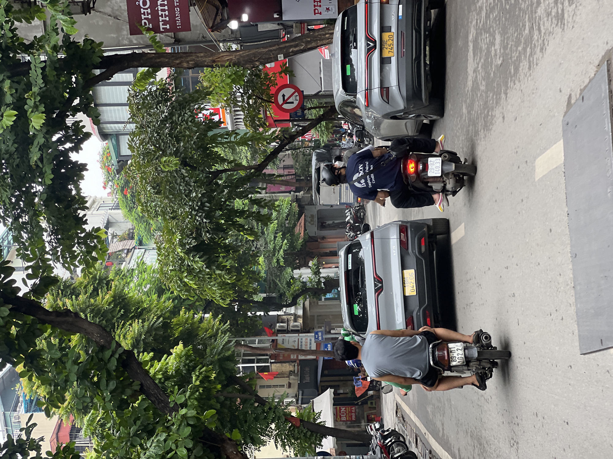 The image captures a street scene in Hà Nội, Vietnam, likely on a daytime with overcast conditions judging by the lighting. The foreground features two motorcycles. On the left, a motorcycle is ridden by a man wearing a grey sleeveless shirt and black shorts, with his back to the camera. On the right, another motorcycle is ridden by a person wearing a dark blue t-shirt with white lettering and a black helmet. Both motorcycles are in motion, driving down a paved road.
Two cars are also prominent in the scene. A silver car, possibly an SUV, is in the middle ground on the left, facing away from the camera. Its license plate reads "89-M1 042.06". To the right of this car, another silver SUV, facing the same direction, has a license plate that says "29E 745.20". Parked motorcycles line the left side of the road.
The environment is urban, with buildings lining both sides of the street. The architecture appears to be a mix of modern and older structures. Trees with dense green foliage overhang the street, casting shadows and contributing to a somewhat lush atmosphere. Signs with Vietnamese text are visible on the buildings, indicating businesses and possibly traffic regulations. One sign in red and white on the right reads "PHỐ THỊNH" and below it "1 HÀNG TRƯỜNG". Another sign shows a red circle with a white horizontal bar and an arrow pointing right, indicating a no-right-turn restriction. The overall impression is of a busy street in a Vietnamese city.