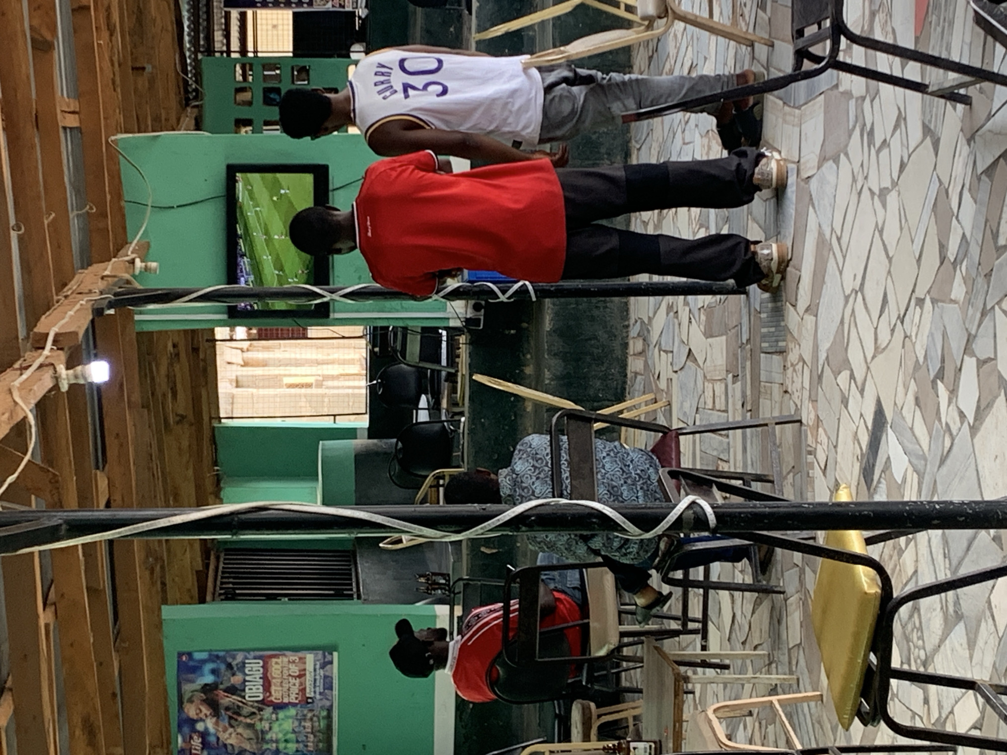 Four men are watching a television inside an open-air bar in New Stadium, Nigeria. The walls of the building are a light green with exposed wooden beams along the roof. A poster for Obiagu is attached to the wall. The men are seated on a variety of metal and wooden chairs arranged facing the television. Two of the men are standing and looking at the television. One man is wearing a red shirt and the other is wearing a white “Curry 30” basketball jersey. The floor is covered in irregularly shaped light-colored tiles.