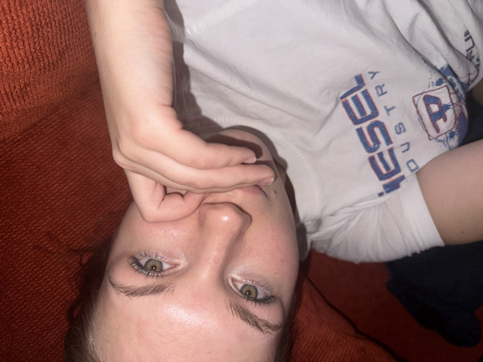 A person lies on a reddish-brown couch, wearing a white t-shirt with blue lettering that reads "Diesel Industry". One hand is raised, partially obscuring the face, while the eyes are open, looking directly at the camera. The skin tone is fair, and the background is dominated by the texture of the couch and a glimpse of what appears to be dark clothing.