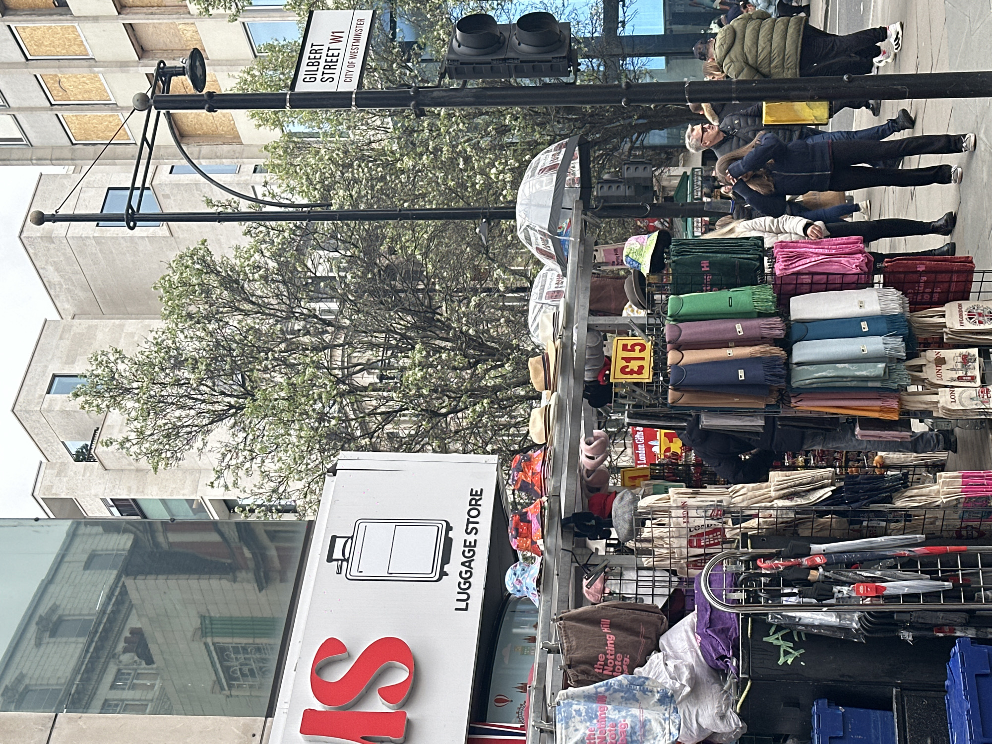 The image shows a street scene, possibly a market, in the City of Westminster, London, United Kingdom. A street sign indicates "Gilbert Street W1." A "Luggage Store" sign with an icon of a suitcase is visible. Displayed on racks are various merchandise, including scarves, hats, umbrellas, and tote bags. Some items are priced at £15. Several people are walking on the sidewalk in the background. The image is vertically oriented. A traffic signal is positioned above the pedestrian crosswalk. Buildings and trees are visible in the background.