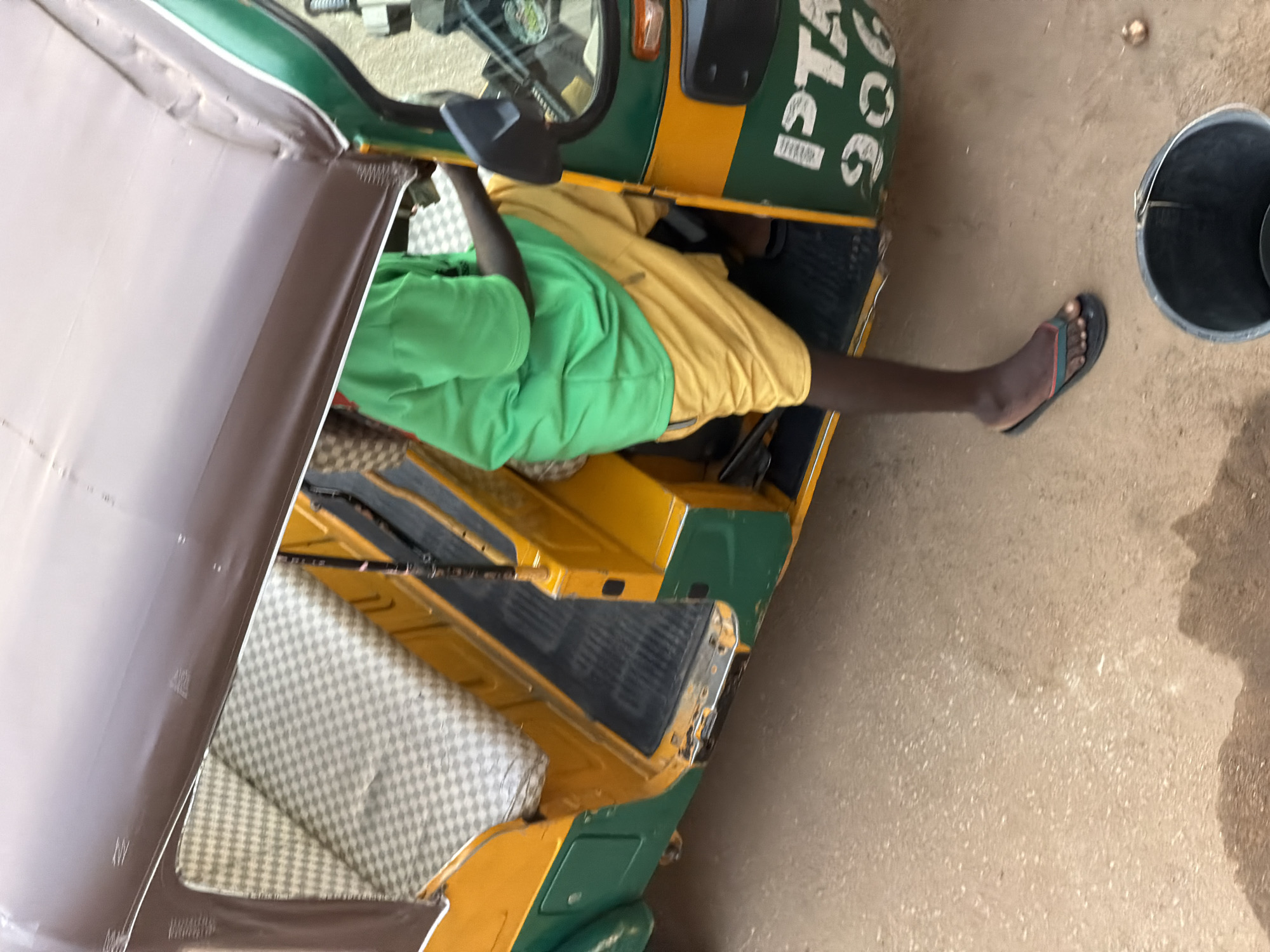 A person wearing a bright green t-shirt and yellow shorts sits inside a yellow and green auto-rickshaw. The auto-rickshaw has a brown roof and checkered seating. The person is wearing black flip-flops and one foot is visible. There is a black bucket on the ground next to the auto-rickshaw. The ground appears to be dirt or sand. White lettering, possibly a license plate or identification number, is visible on the side of the auto-rickshaw. The image seems to be taken from a slightly elevated angle. The scene is consistent with the environment found in Bukuru, Nigeria, where auto-rickshaws are a common mode of transportation.