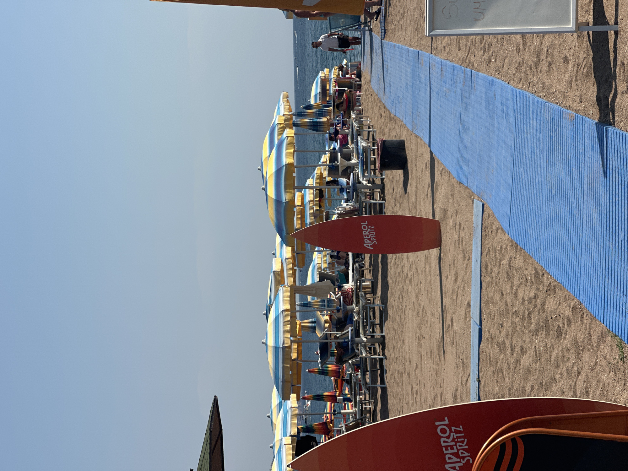 The image depicts a sandy beach scene in Boreti, Montenegro, on a clear, sunny day. A blue mat pathway runs across the sand, leading towards the ocean. On the left side of the image, a red surfboard with "APEROL SPRITZ" branding is prominently displayed. Further into the scene, rows of beach chairs and umbrellas, predominantly yellow and blue striped, are set up on the sand. Some individuals are visible relaxing on the chairs, while others are seen walking or standing. The sea is calm and blue in the background, with a few boats visible on the horizon. A man in a white shirt and dark shorts walks along the beach near the pathway, and a white board on a stand is positioned to the right of the pathway. The environment suggests a typical beach resort atmosphere.