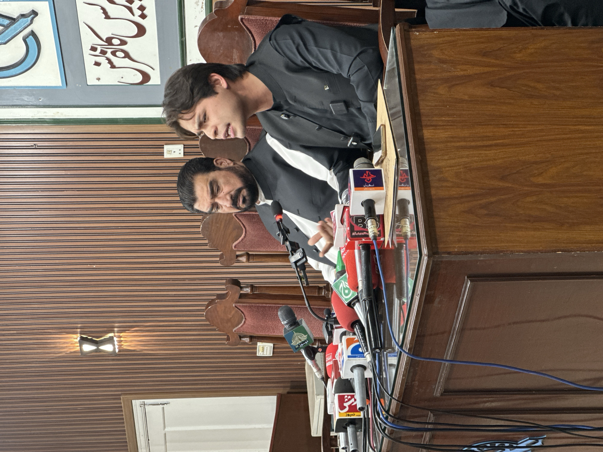 The image depicts an indoor scene resembling a press conference or media briefing. Two men are seated behind a dark wooden table or podium, facing a cluster of microphones.
The man on the left (when the image is correctly oriented) appears younger, with dark hair, and is wearing a black shalwar kameez-style outfit with a matching waistcoat. He is leaning forward slightly with his mouth open, suggesting he is speaking or listening intently. The man on the right is older, with a beard, and is also dressed in a black shalwar kameez with a white shirt visible underneath. He looks downwards, possibly at notes or the microphones. A third person's arm and torso are partially visible in the background, to the right of the younger man.
In front of the men is an array of microphones, bearing logos of various news organizations. Visible text on the microphone flags includes "NEWS HD", "سچرین" (Sachrain), "سحر" (Sahar), and "نیوز" (News), indicating a diverse media presence. Numerous audio cables, predominantly blue and black, snake across the table.
The background features a wall decorated with large panels of calligraphy in what appears to be Arabic or Persian script, rendered in blue and brown on a white background. One piece of calligraphy looks like the word "مذاکرات" (Muzakerat), meaning negotiations or discussions. The wall or ceiling has a distinct pattern of horizontal dark stripes on a lighter brown surface, with a small, square-shaped light fixture emitting light. The room is furnished with dark wooden chairs with upholstered backs. The overall atmosphere suggests a formal public announcement or statement in an unknown location.